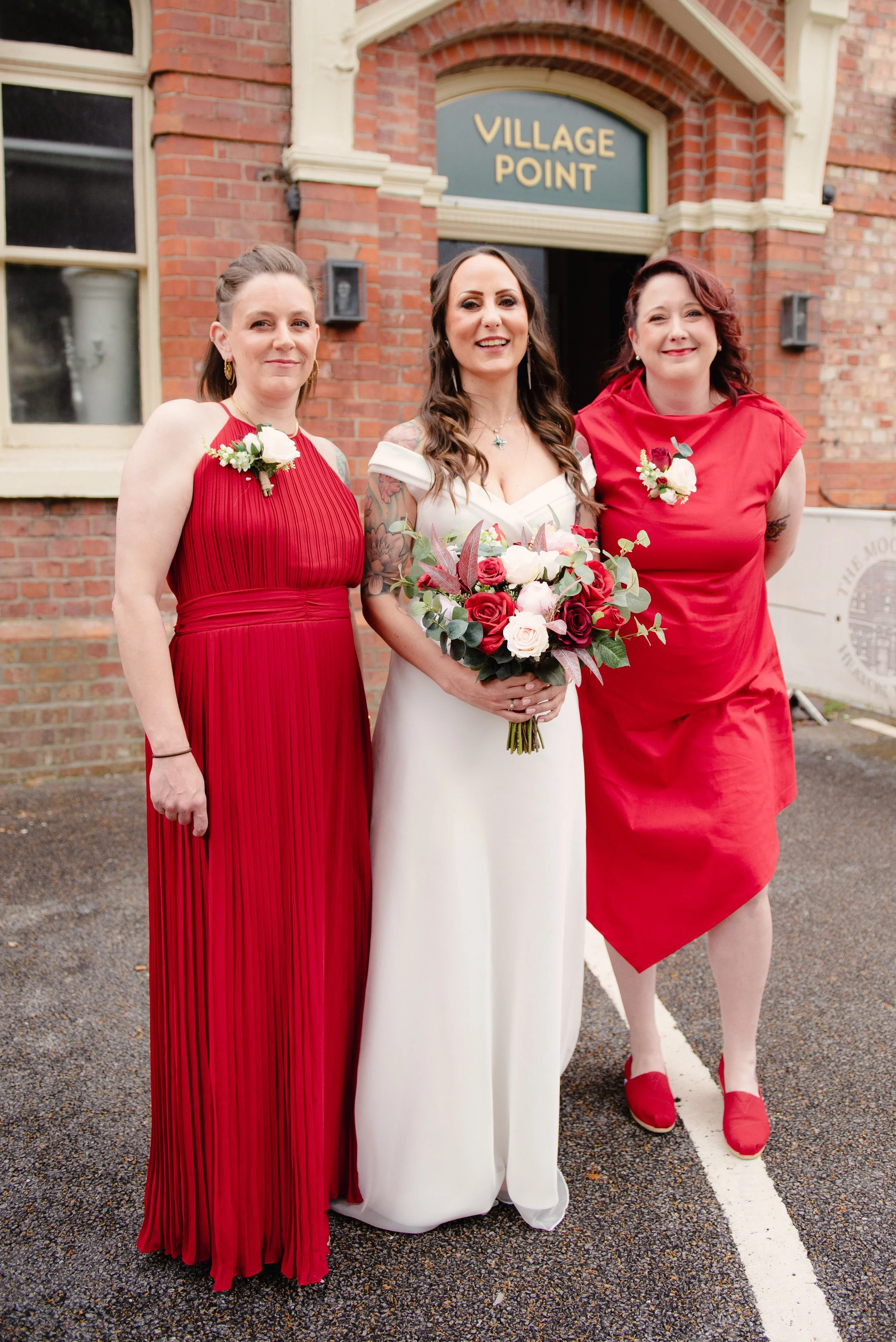 Three women dressed in red and white, standing outside a brick building with a sign that says 'Village Point'. The woman in the middle is wearing a white wedding dress and holding a bouquet of pink, white, and red flowers. The woman on the left and t