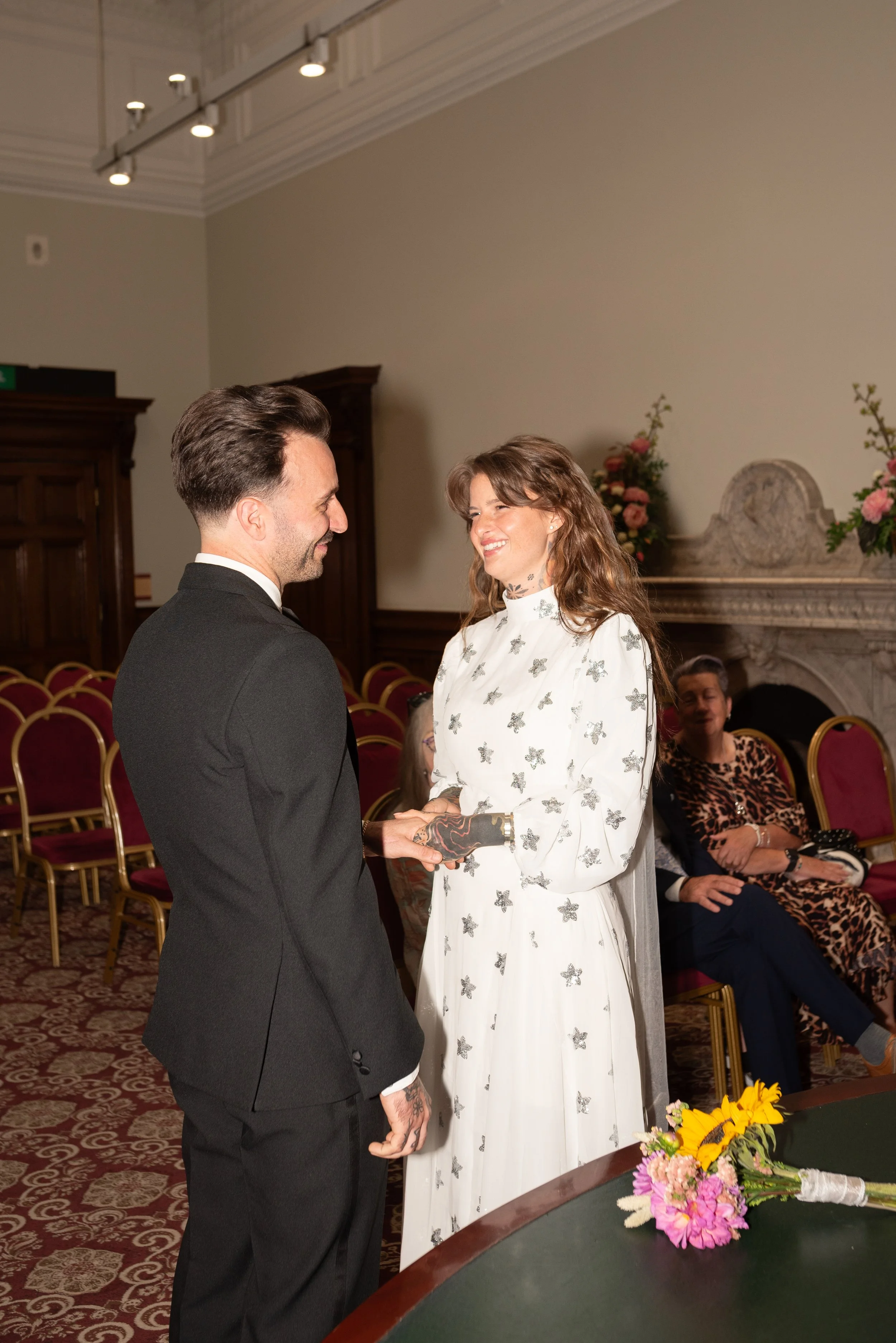 A couple is exchanging rings at their wedding ceremony, with friends observing in the background.