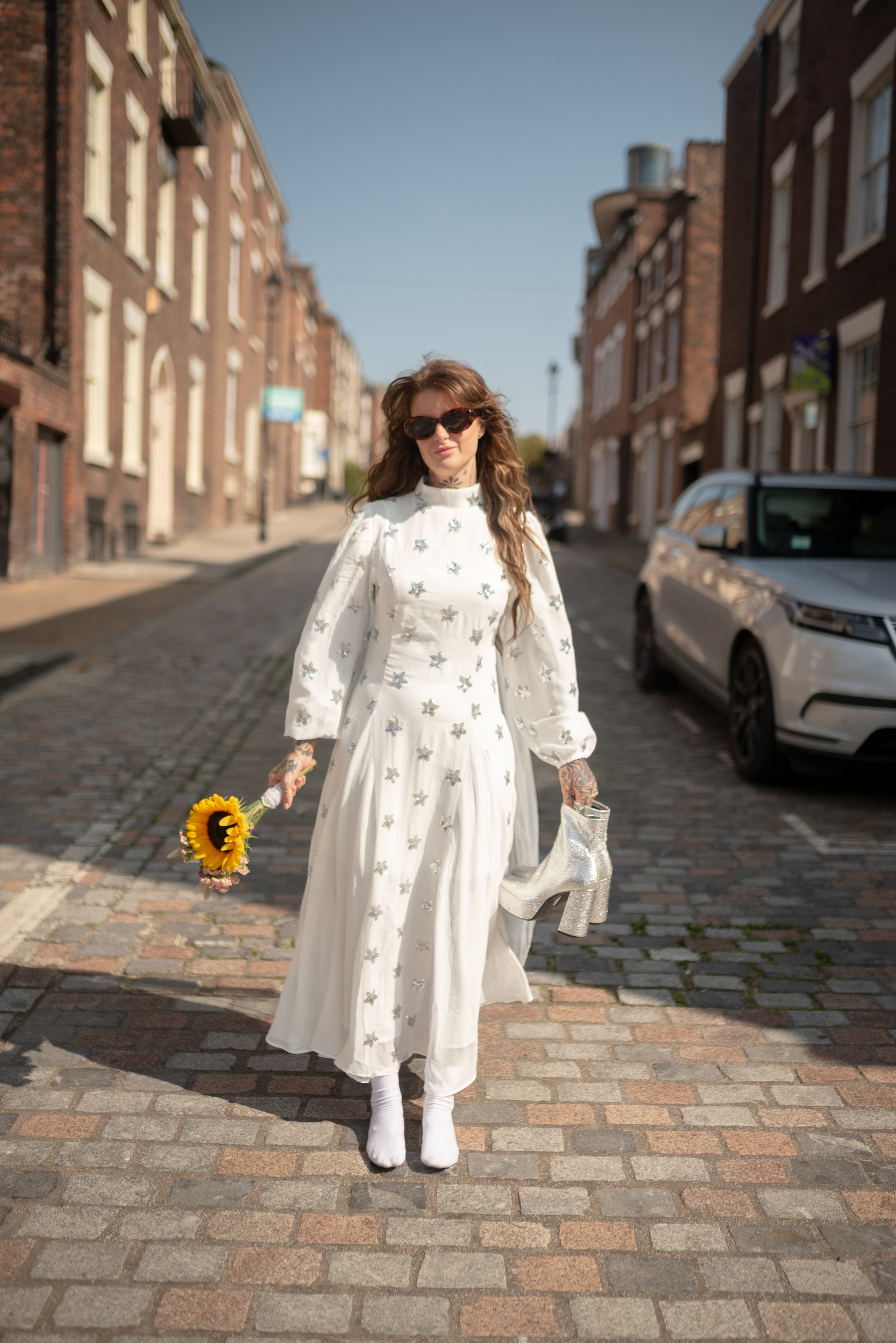 A woman with long wavy brown hair, wearing sunglasses and a long white dress decorated with small silver embellishments, stands on a cobblestone street holding a sunflower bouquet in one hand and silver glitter high-heeled boots in the other.