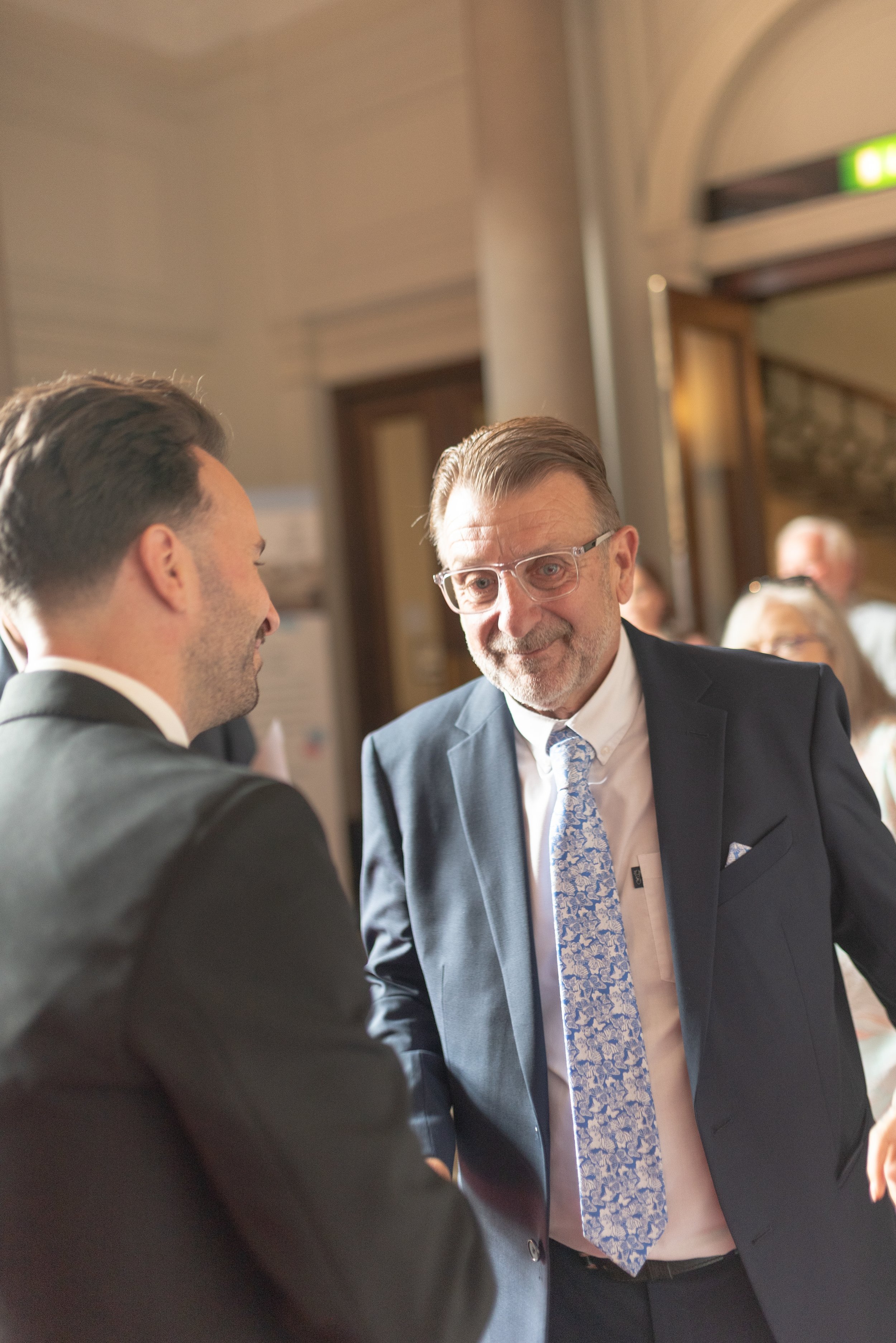 Two men in suits conversing at a formal event, one with glasses and a blue patterned tie, the other with short dark hair, in an elegant indoor setting.