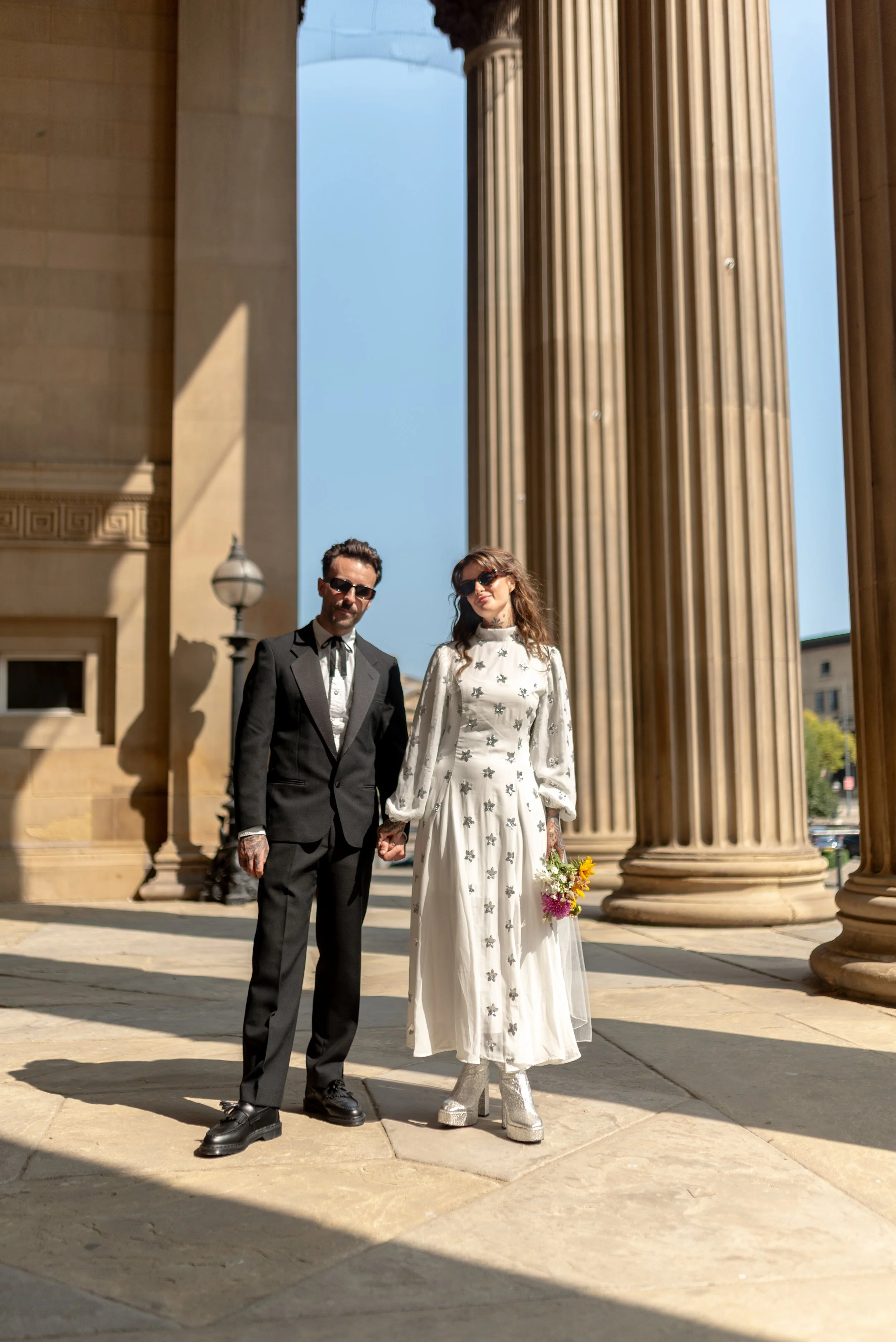 A couple standing hand-in-hand in front of large classical columns on a sunny day. The man is dressed in a black suit and tie, wearing sunglasses. The woman is dressed in a vintage style white dress with floral embroidery and shiny platform boots, ho