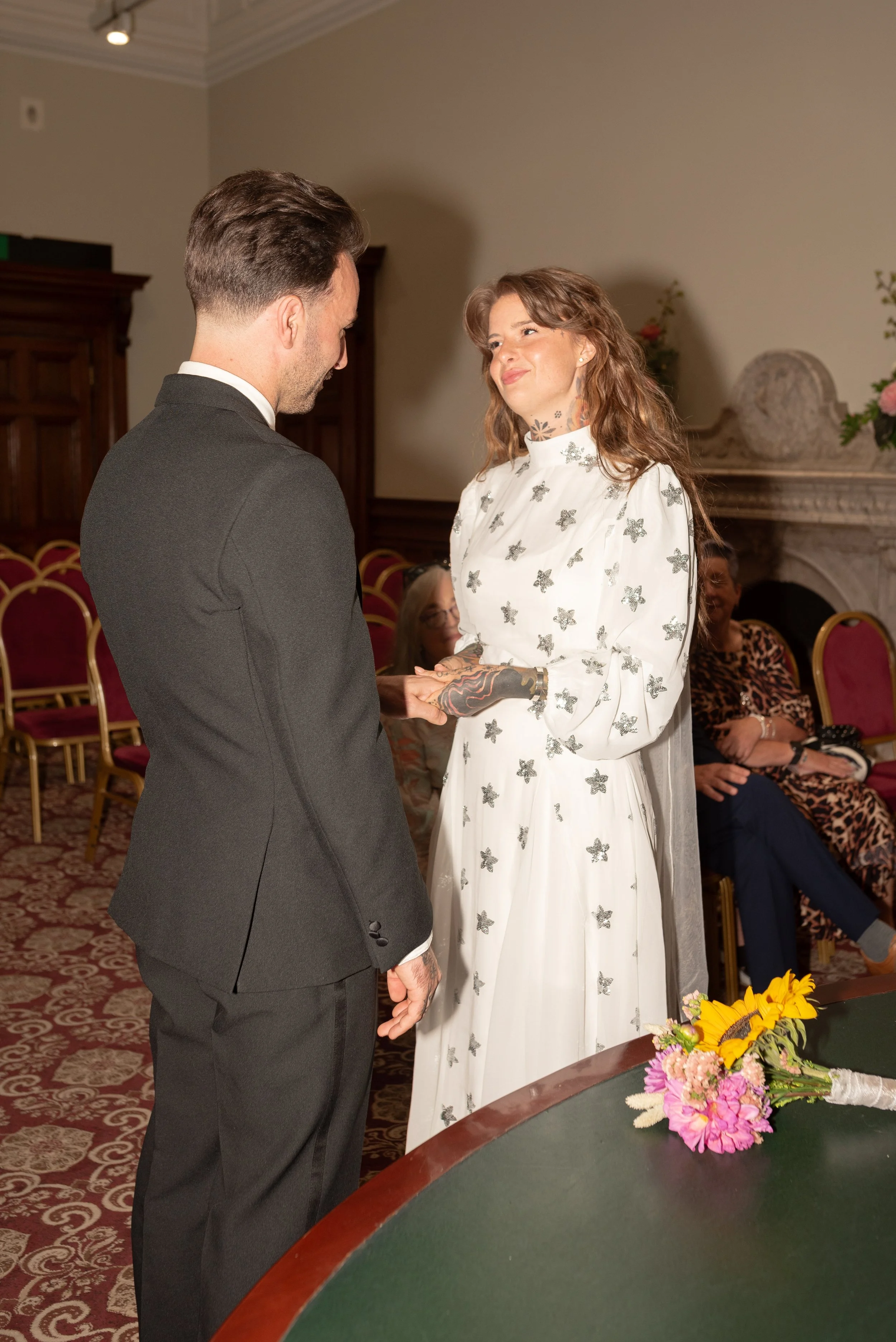 A woman in a white dress with metallic butterfly patterns is holding hands with a man in a black suit during a wedding ceremony. They are standing indoors in a room with red chairs and a floral arrangement on a table.