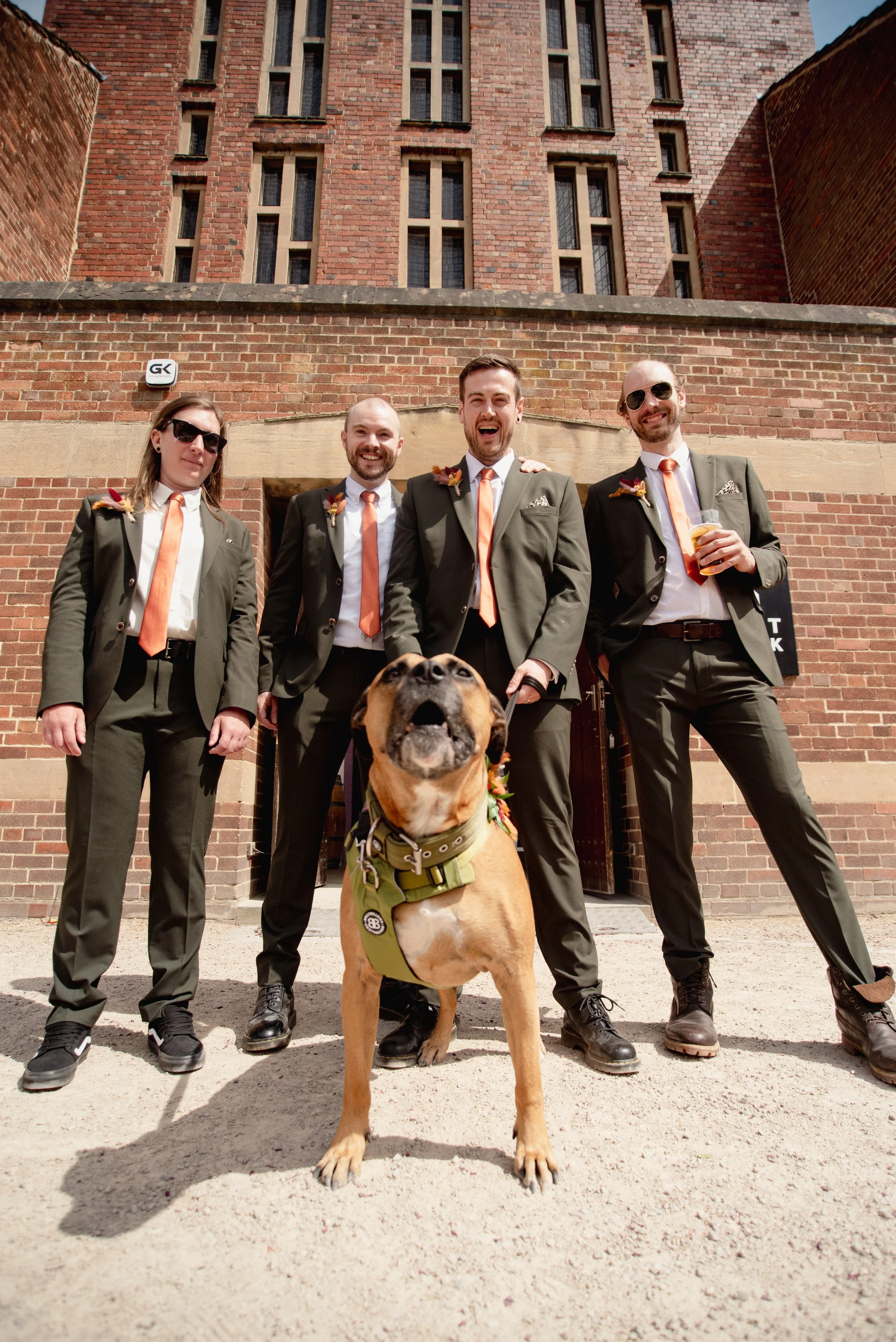 Four men and a woman dressed in formal suits with orange ties, standing together outside in front of a brick building. They are smiling and appear happy, with one holding a drink. There is a large dog in front of them, sitting and looking upwards.