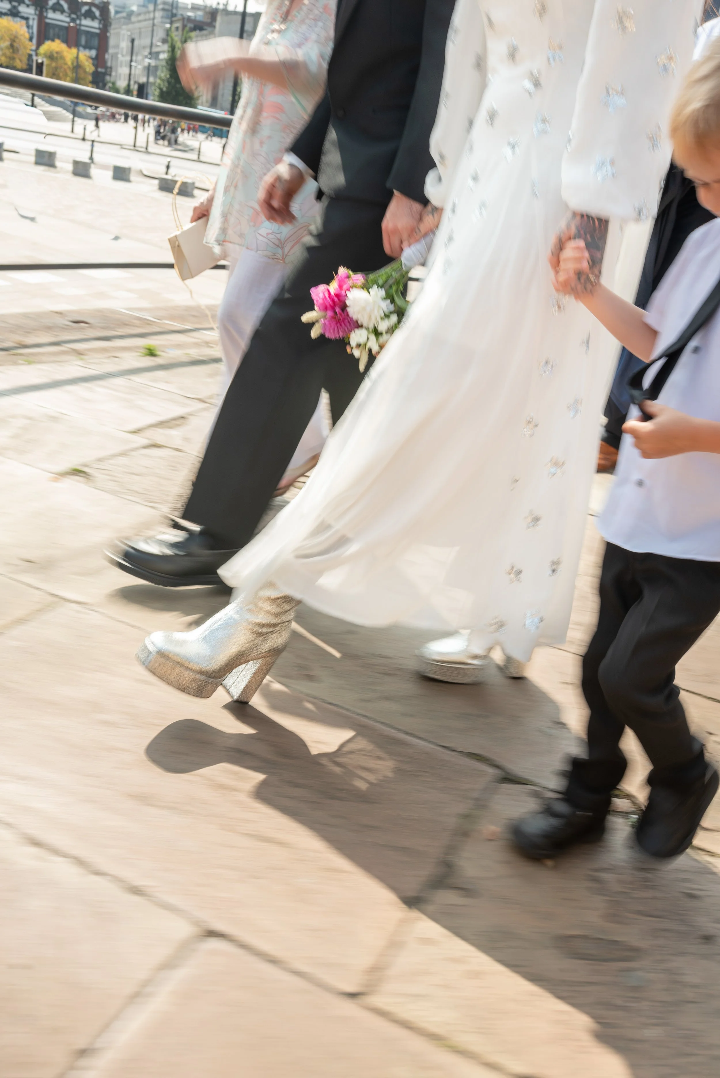 Close-up of people walking outdoors, one person holding a bouquet of pink and white flowers, wearing a white dress with shiny silver high heels, another person wearing black shoes and black pants, and a child in a white shirt holding an adult's hand.