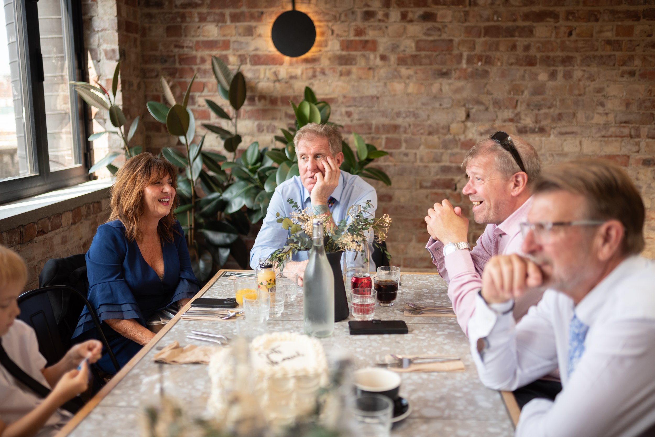 A group of adults and children sitting at a dining table in a restaurant with a brick wall and large window in the background, enjoying each other's company.