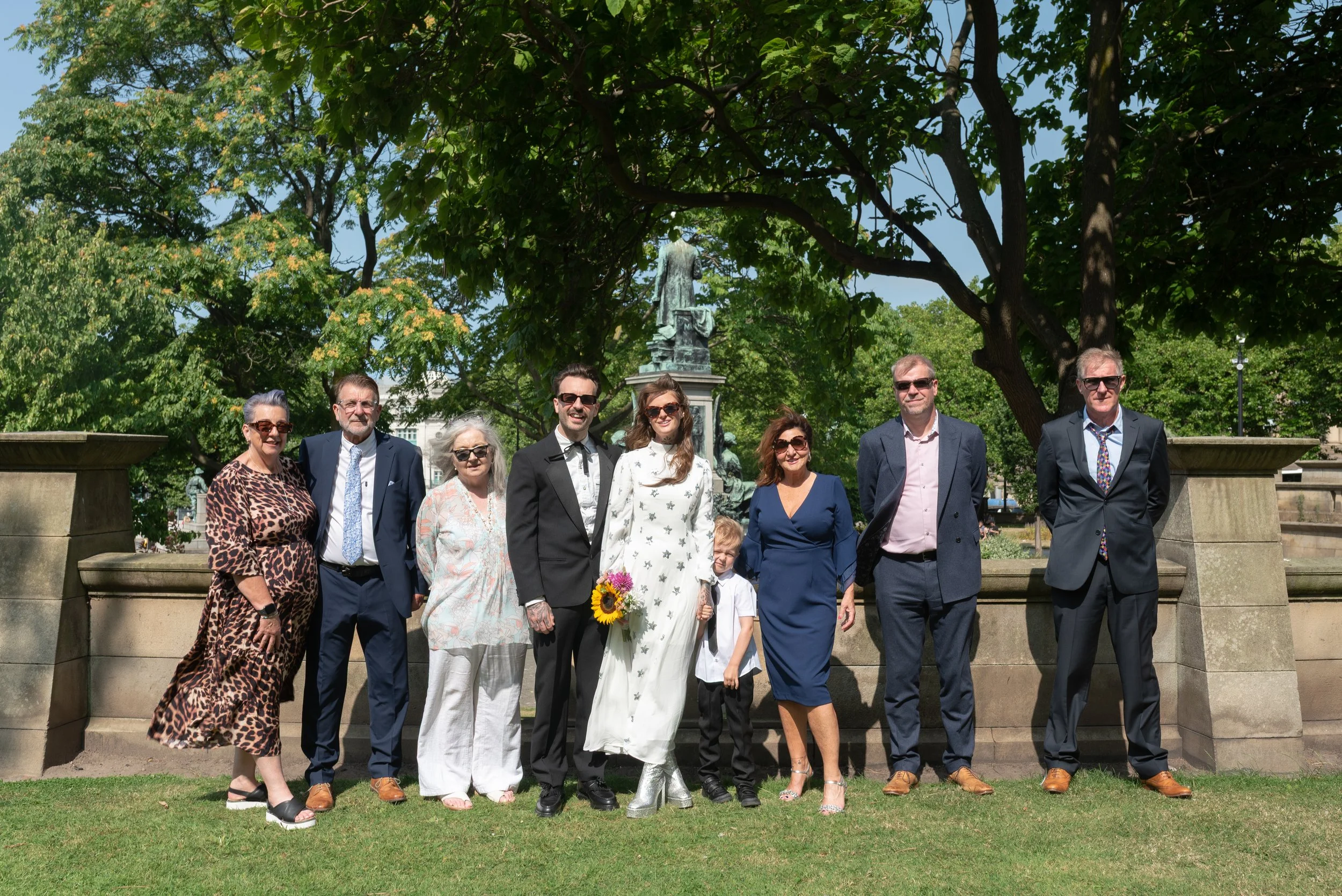 Group of ten people, including adults and a child, standing outdoors on grass in front of a stone wall with a bronze statue and a large tree.