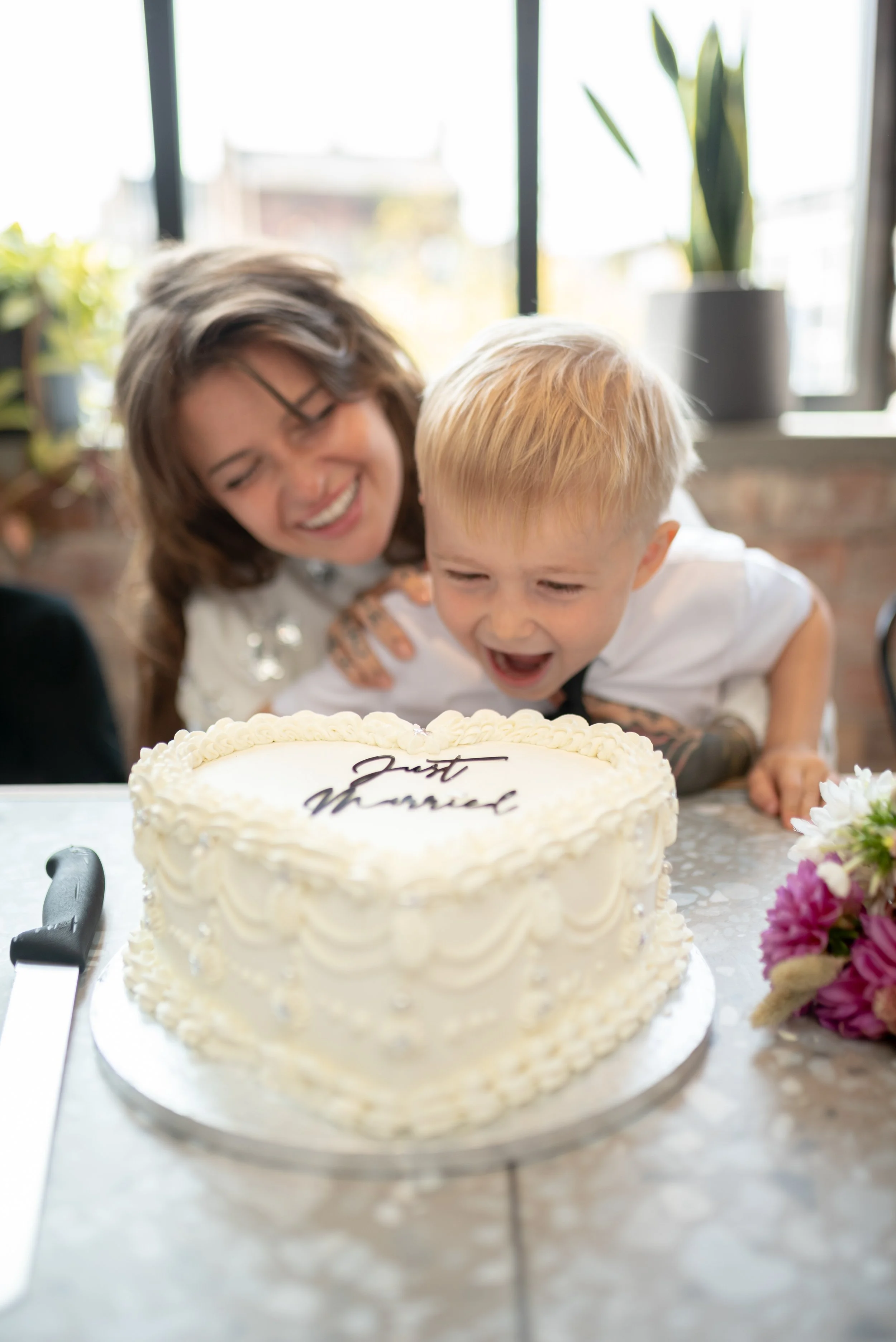 A young boy and a woman, likely his mother, are leaning over a heart-shaped white birthday cake, smiling and celebrating. The cake has the words 'Just Married' written on it.