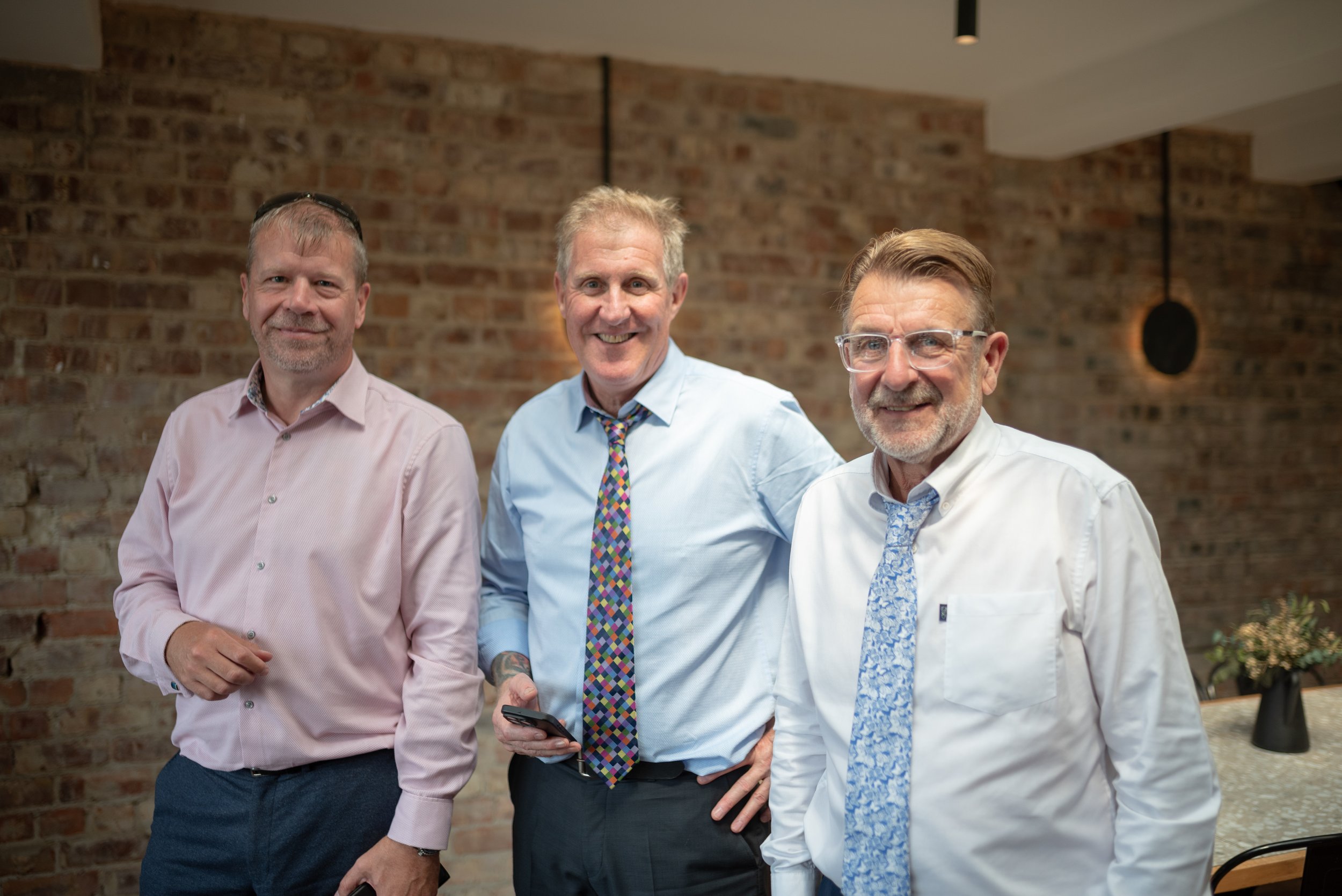 Three middle-aged men dressed in business casual attire standing and smiling indoors with a brick wall background.
