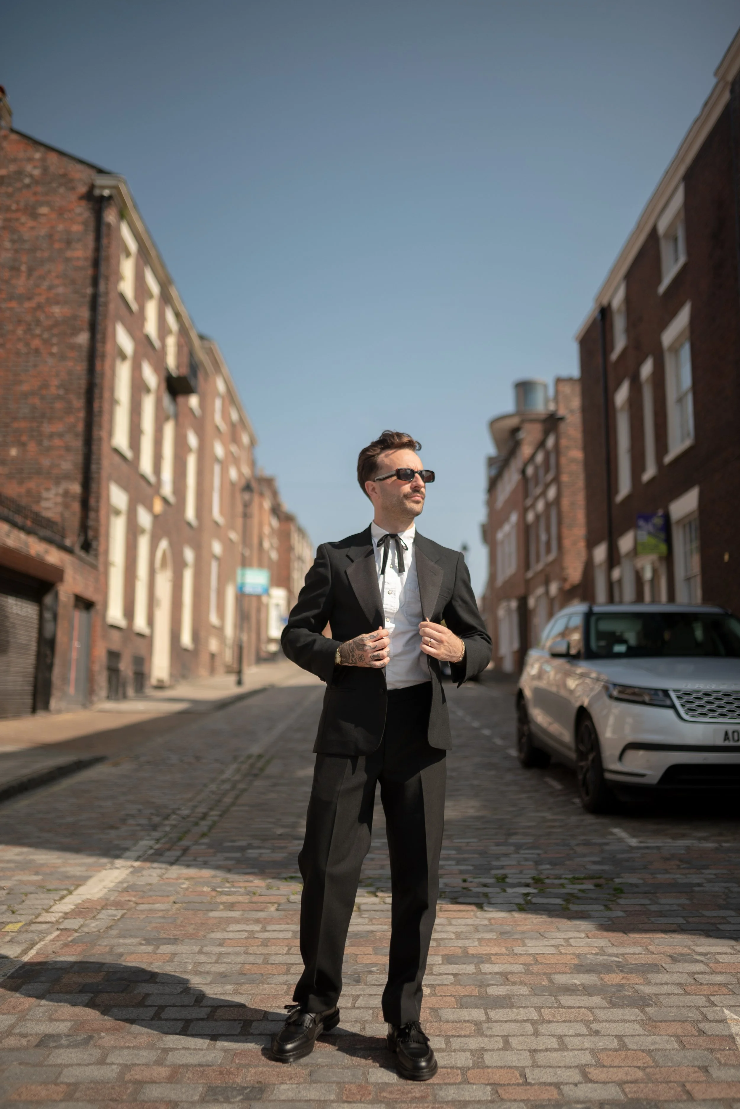 Man in a black suit, white shirt, and sunglasses standing on a cobblestone street with brick buildings and a silver car in the background.