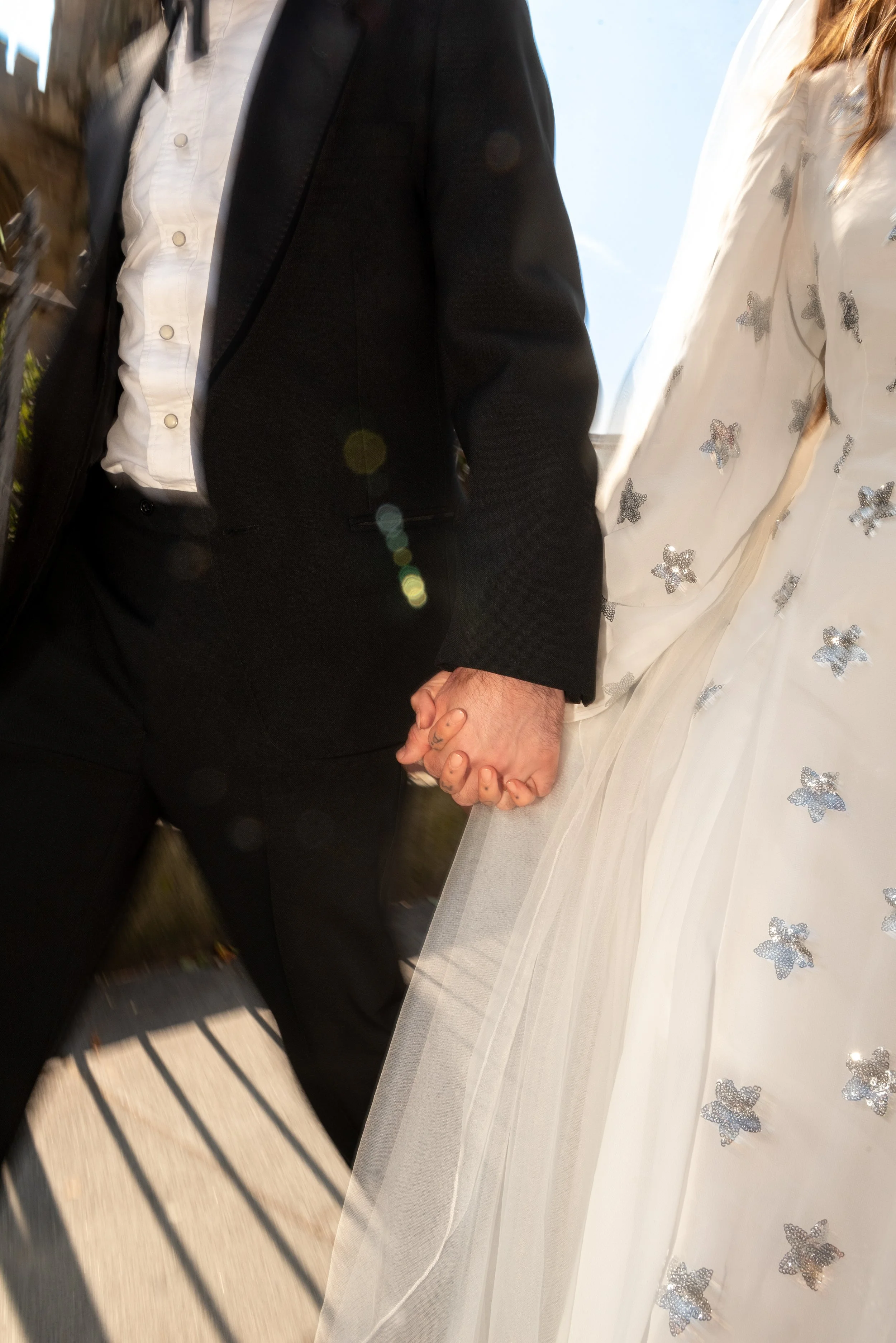 Close-up of a couple holding hands, with one person dressed in a black suit and the other in a white dress with butterfly patterns.