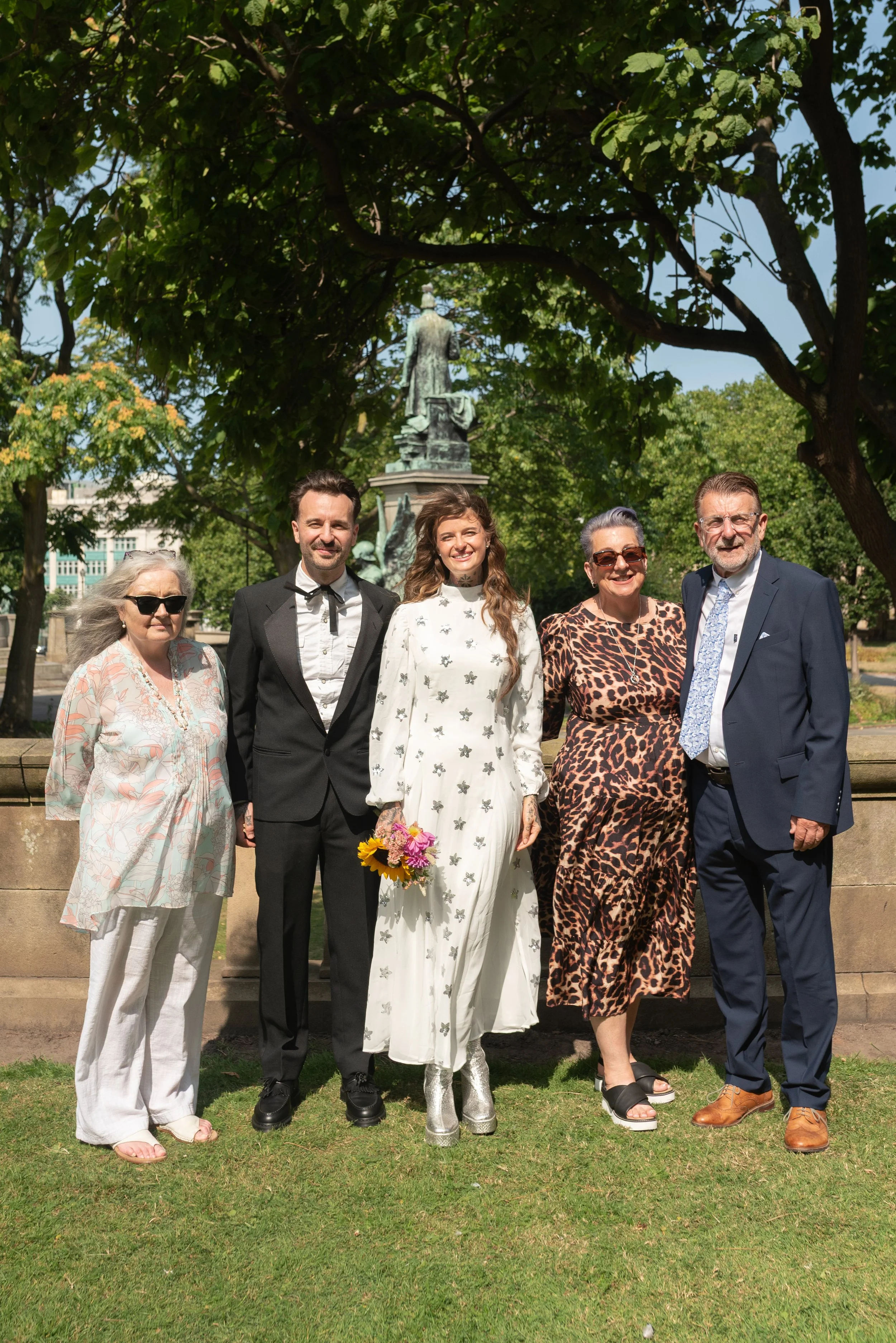 Group of six adults standing outdoors in front of a tree and a statue, dressed in semi-formal attire, smiling for a photo.