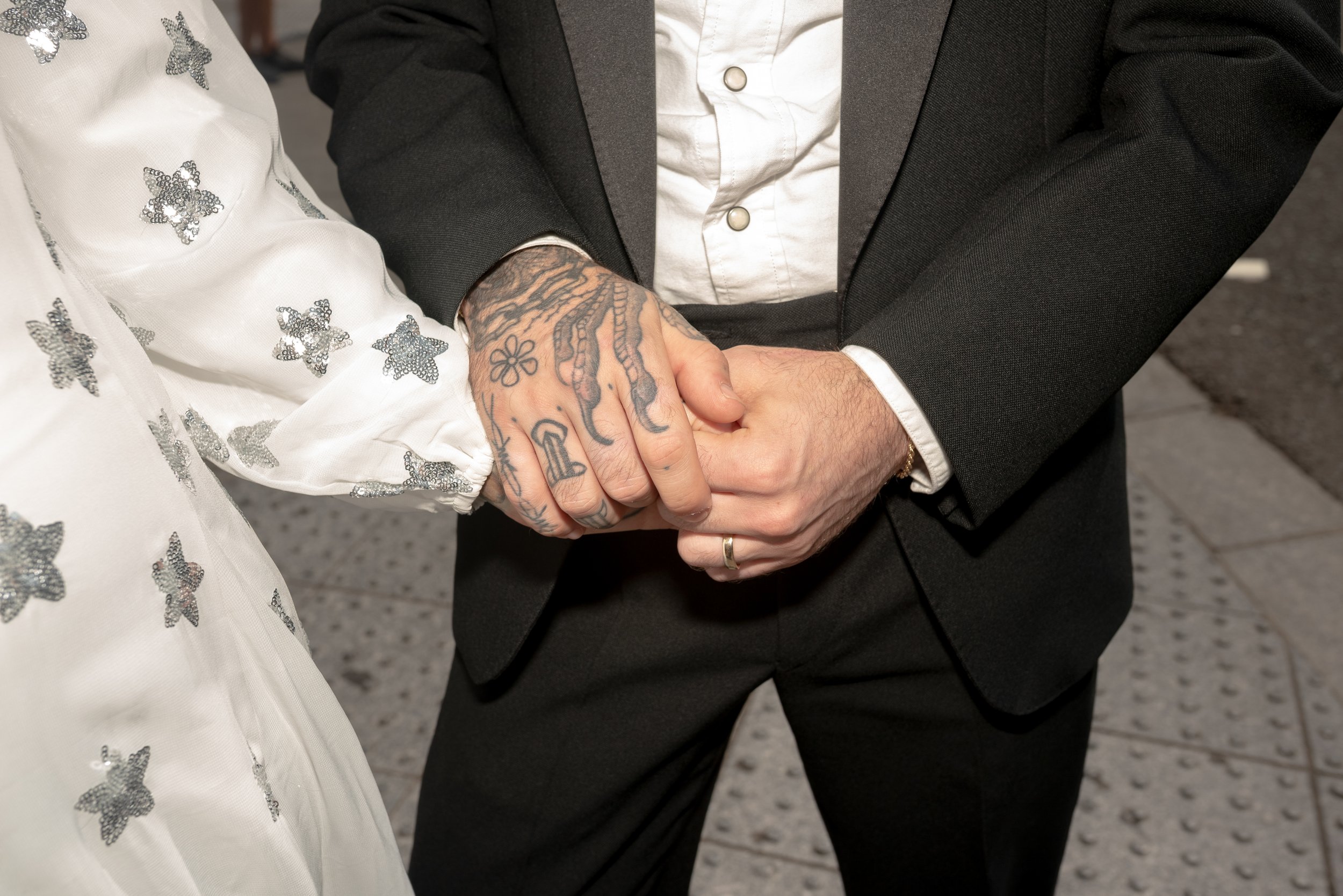 Close-up of a couple holding hands, with the person on the left wearing a white dress with silver star patterns and the person on the right wearing a black suit. The person on the right has tattoos on their hand and wrist.