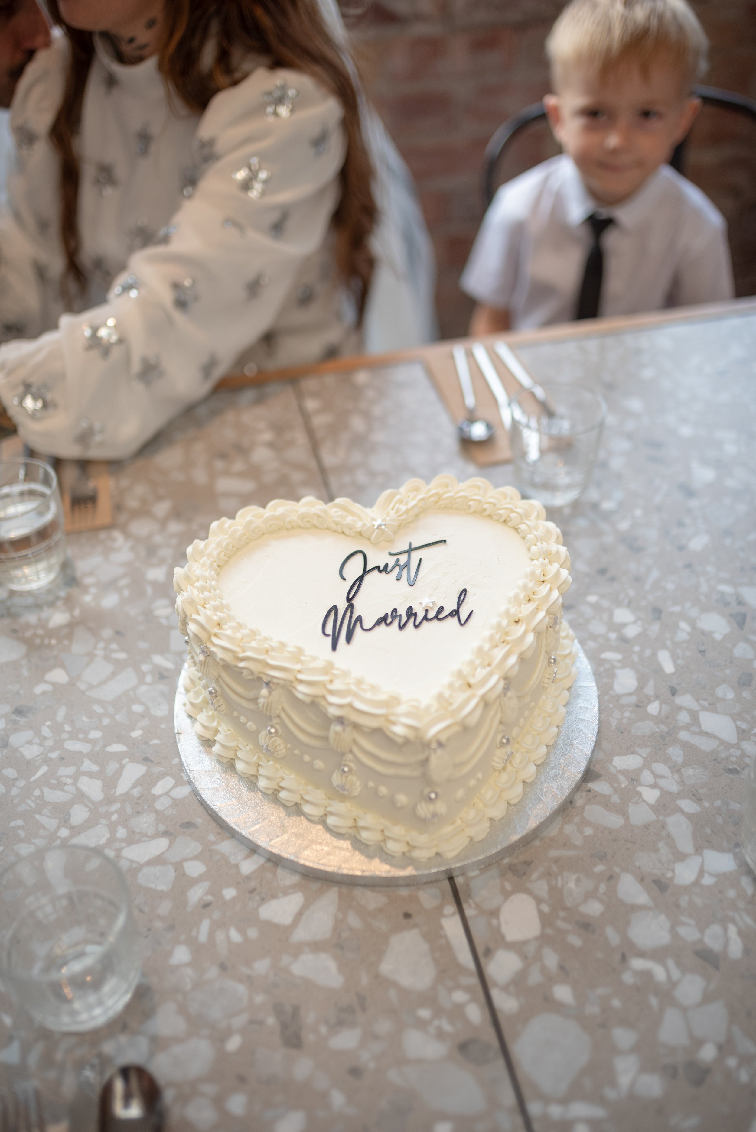Heart-shaped wedding cake with white icing and decorative pearls, with 'Just Married' written on top, placed on a table with forks and glasses, with two children sitting nearby.