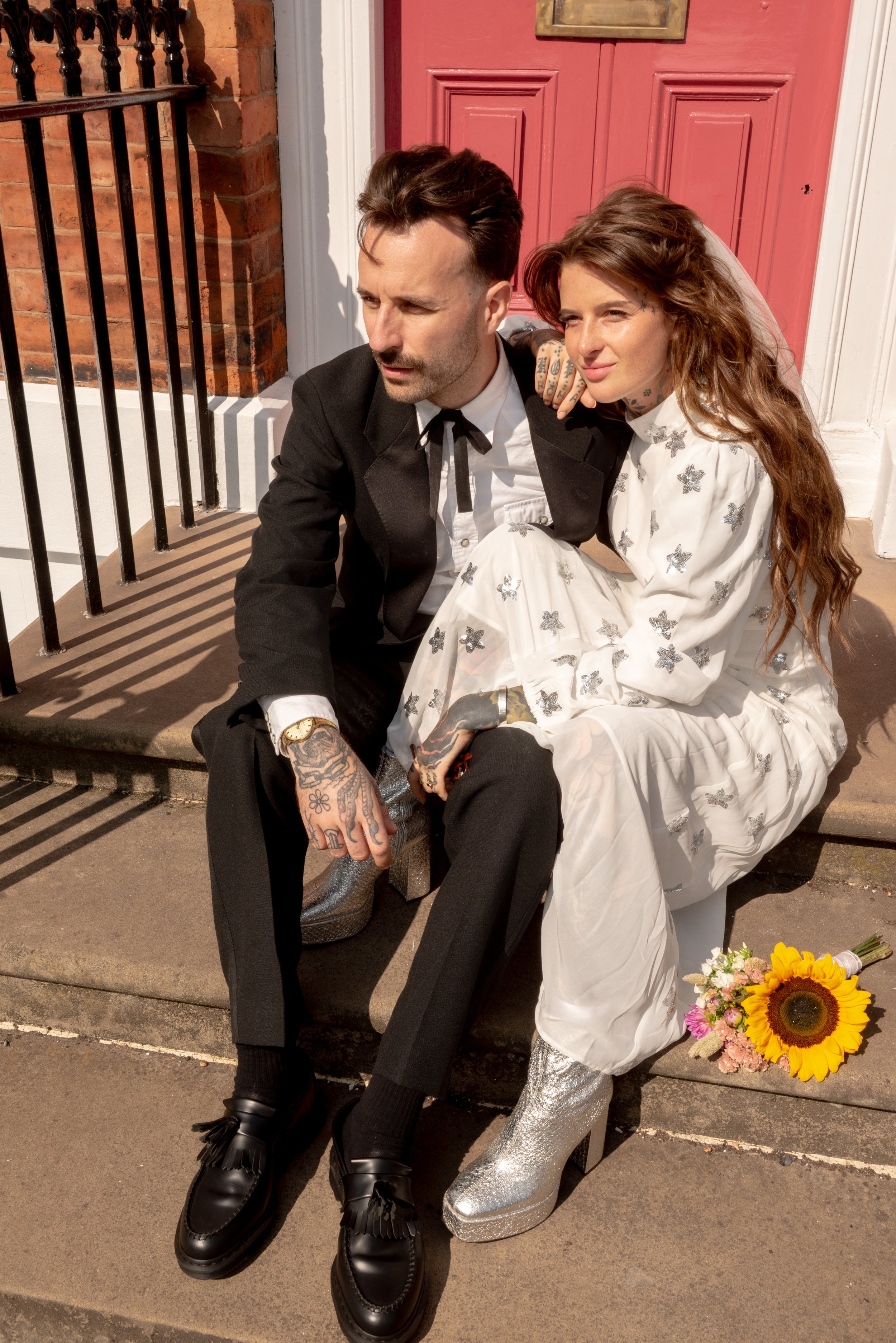 A man and a woman sitting on the sidewalk in front of a red door. The man is dressed in a black suit, white shirt, and black shoes, with tattoos on his hands. The woman is wearing a white dress with butterflies, sparkly silver boots, and has long, wa