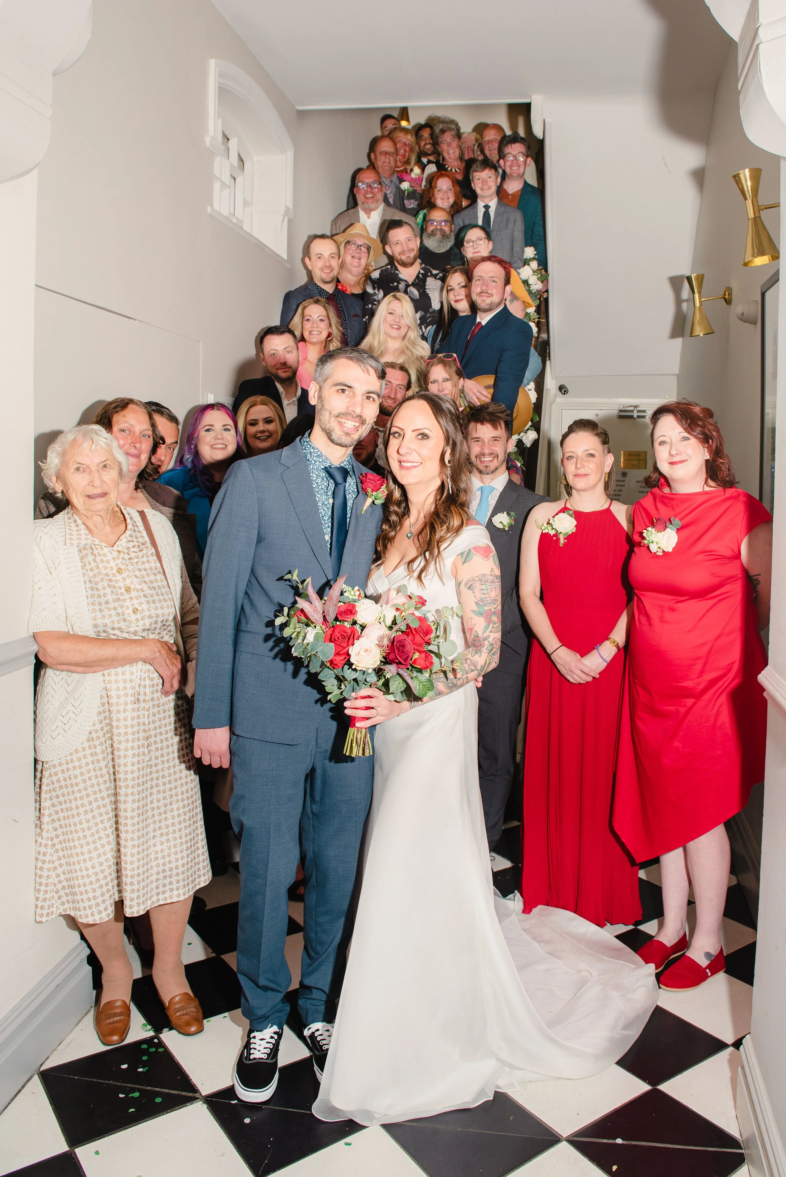 A wedding party group photo indoors showing a bride and groom in front, surrounded by family and friends on a staircase and hallway.