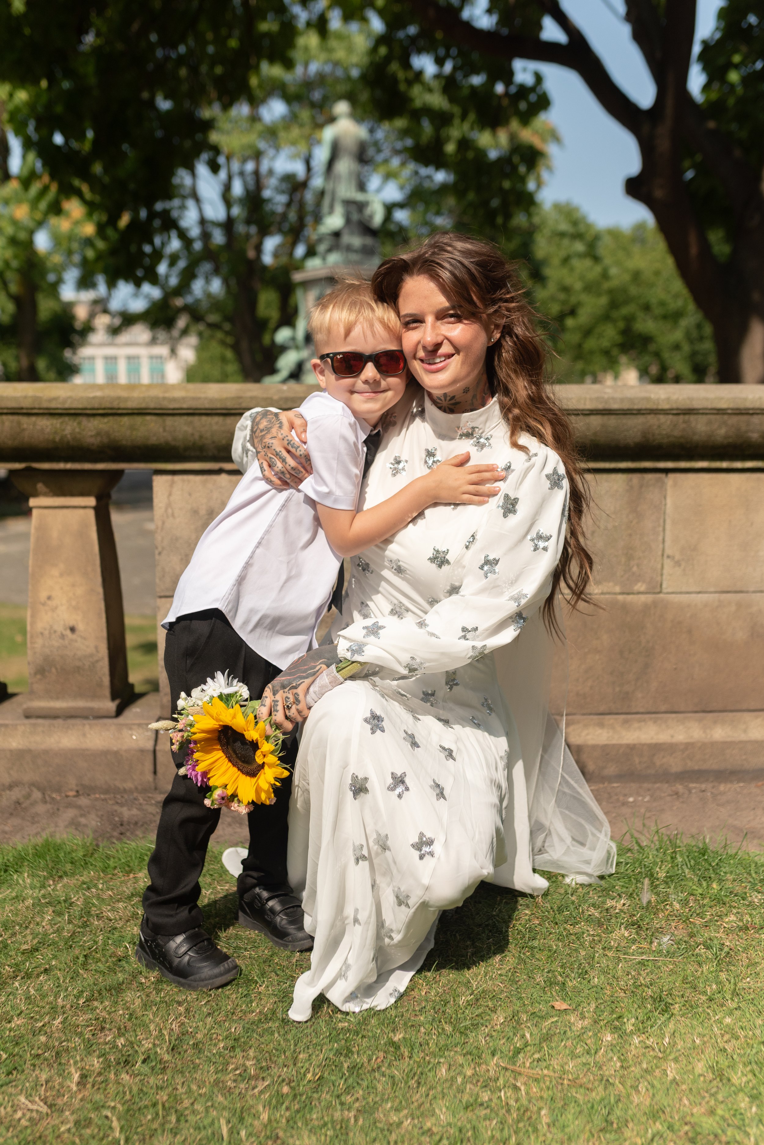 A woman in a white dress with butterfly patterns kneeling on grass, holding a sunflower, hugging a young boy wearing sunglasses and a white shirt, in a park with trees and a statue in the background.