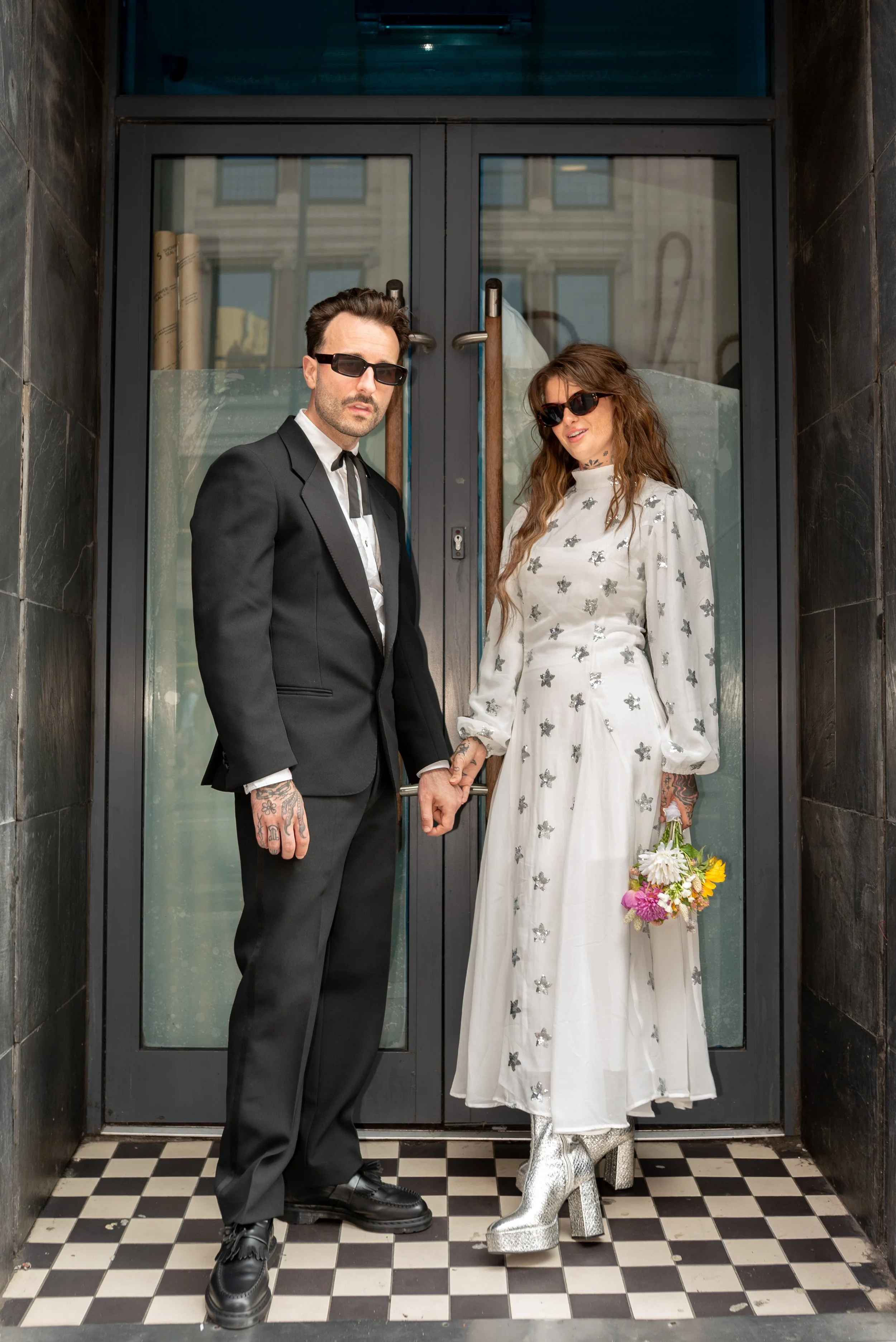 A man and woman dressed in stylish clothing, holding hands, standing outside a building with glass doors, with the man wearing a black suit and sunglasses, and the woman in a white dress with silver embellishments, silver high-heeled boots, and holdi