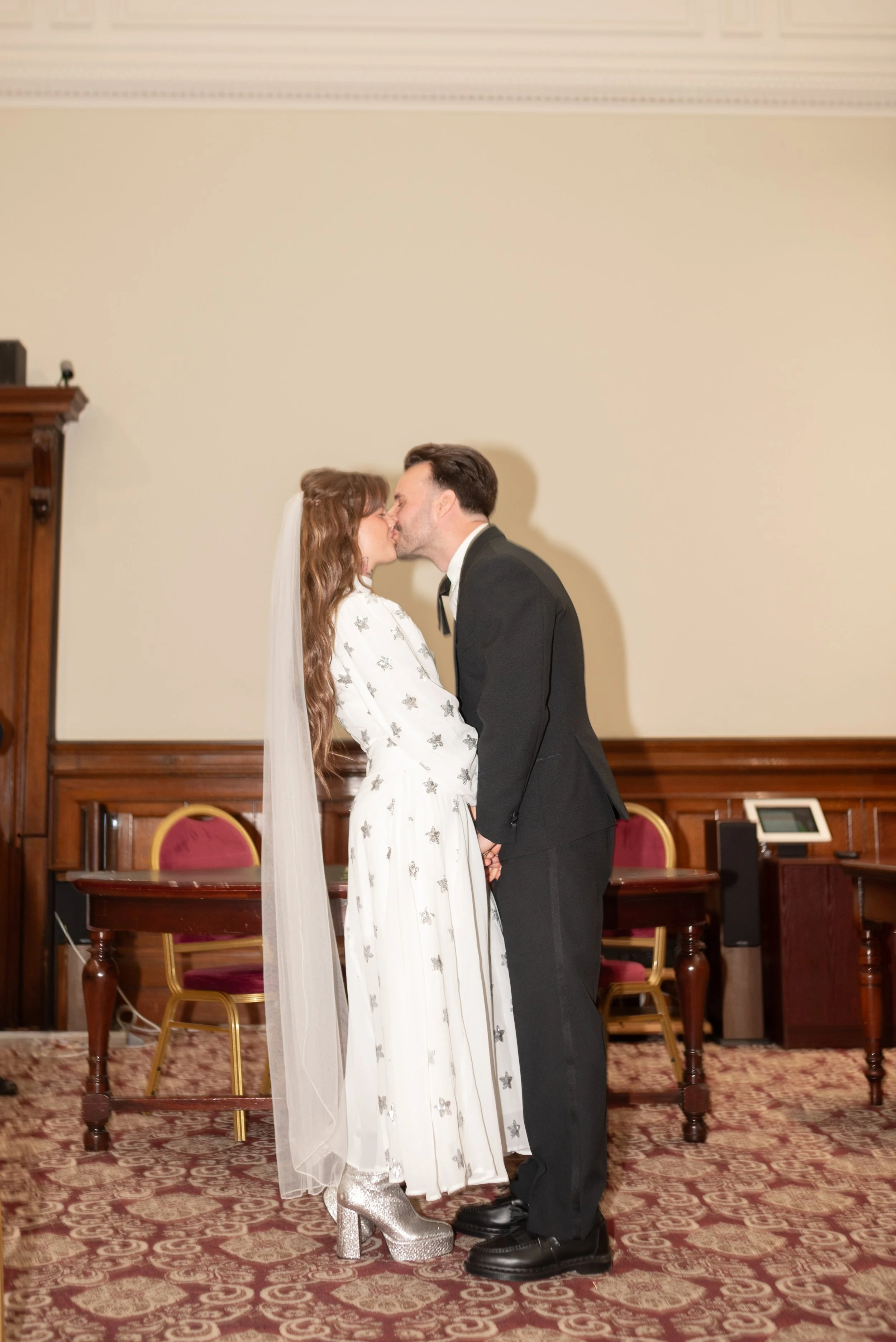 A bride and groom sharing a kiss in a wedding ceremony, standing on a patterned carpeted floor in a room with wooden paneling and red upholstered chairs.