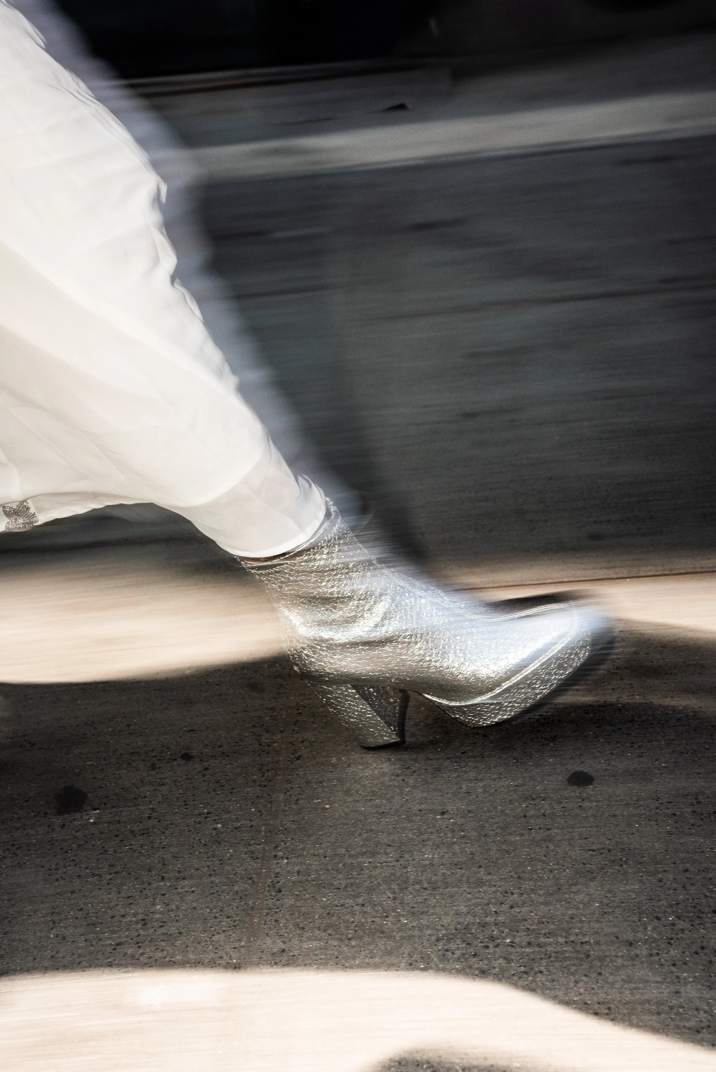 Close-up of a person's leg and foot in a glittery high-heeled shoe, walking on a dark asphalt street at night.