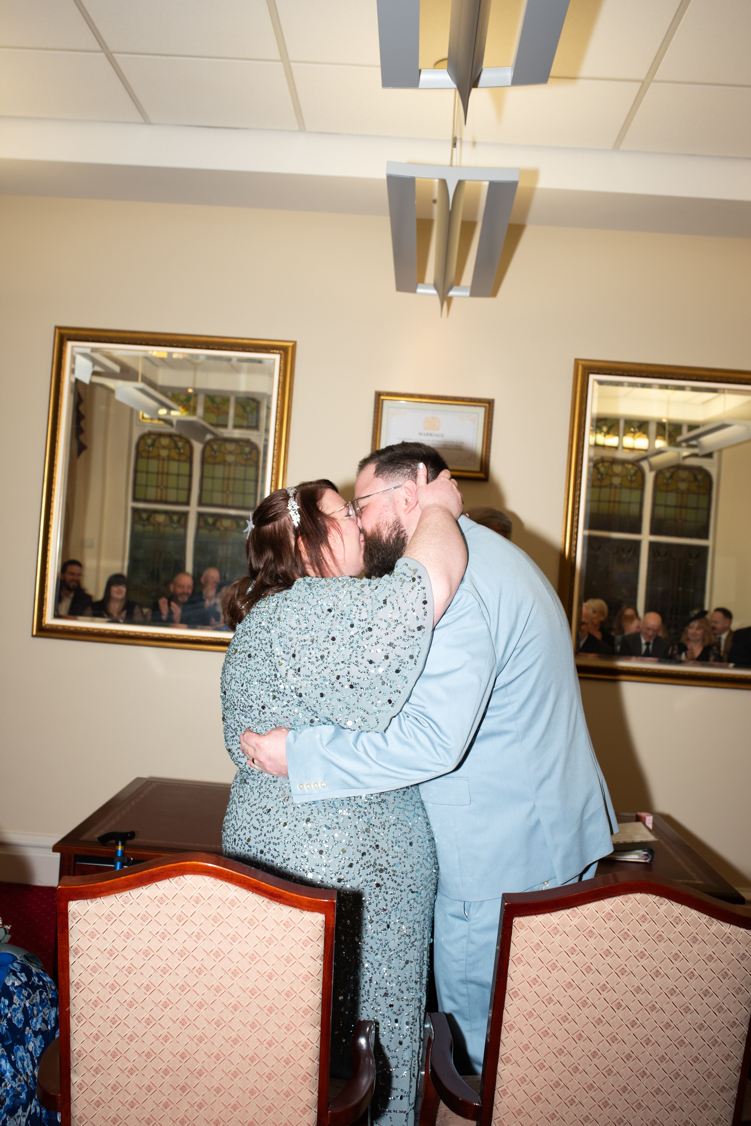A couple shares a kiss during a wedding ceremony, with a background of friends and family visible through large mirrors.