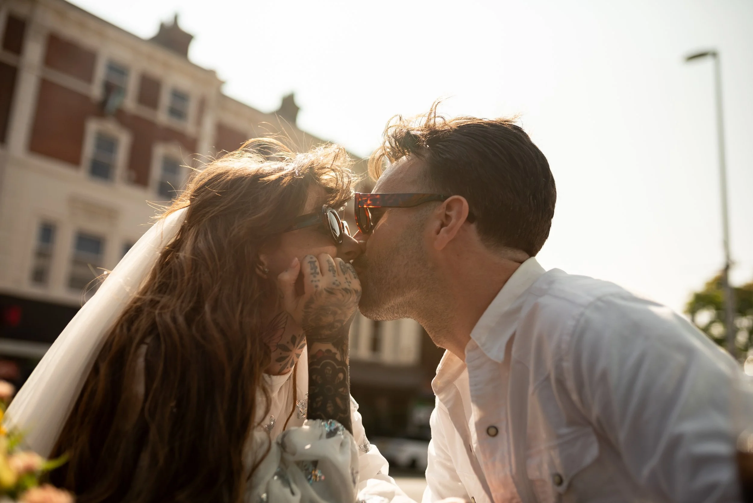 A couple sharing a kiss outdoors during sunset, with buildings and a streetlamp in the background.