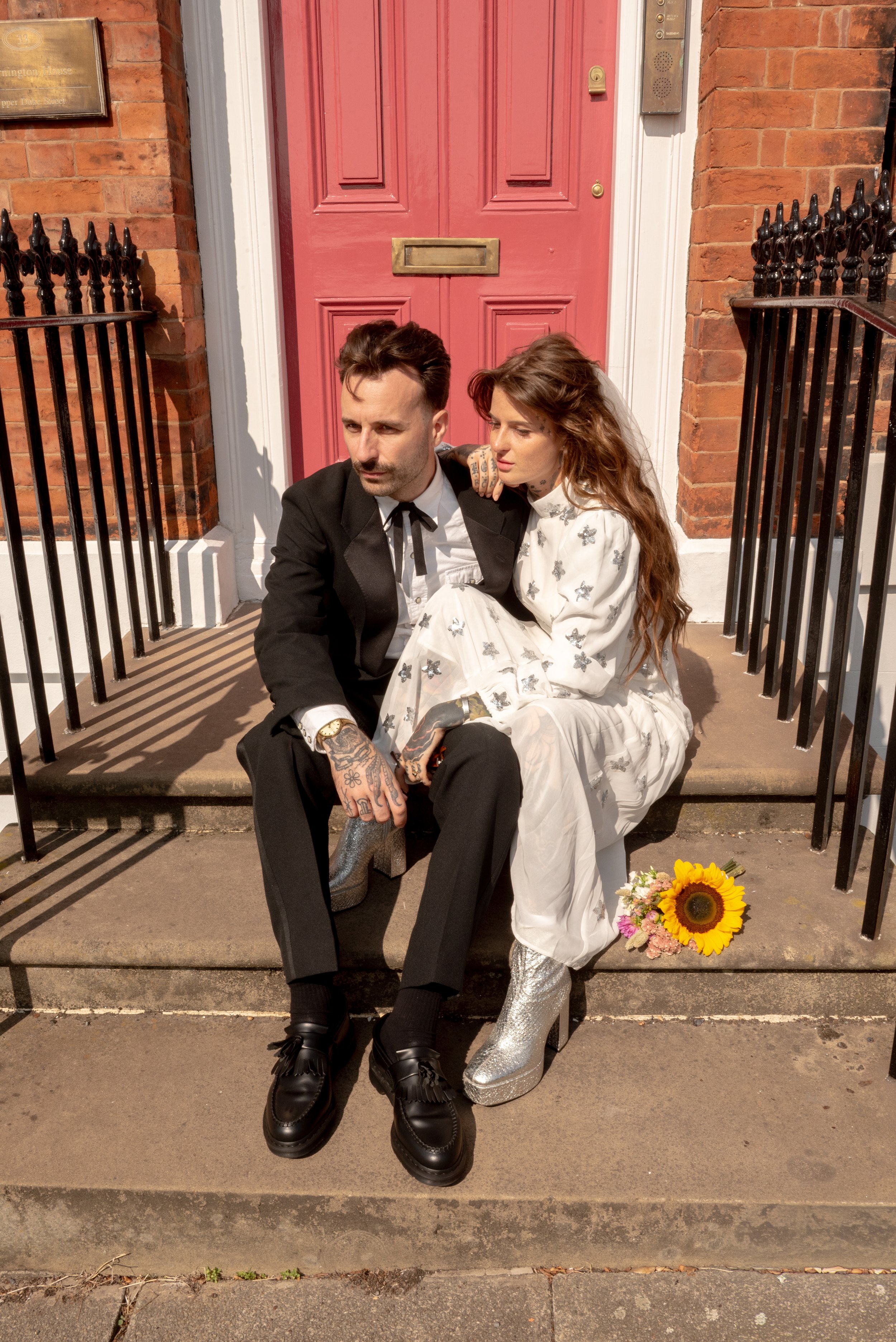 A man and a woman sitting on the steps outside a house with a red door. The man wears a black suit with a white shirt and black tie, and has tattoos on his hands. The woman wears a white dress with black butterflies and silver boots, holding a sunflo