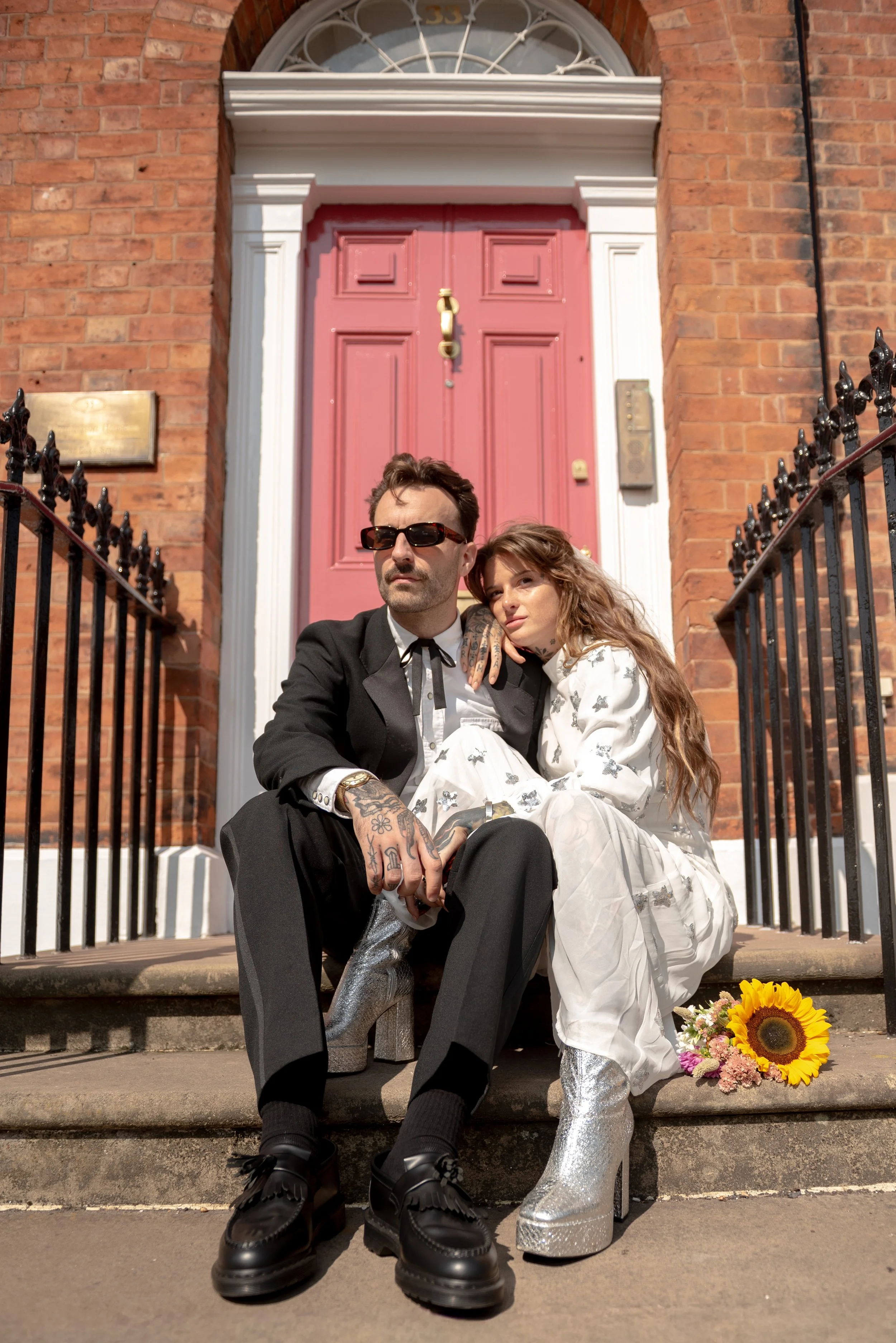 A man and woman sit on front steps of a brick house in front of a pink door. The man wears sunglasses, a black suit, and shiny black shoes. The woman has long wavy hair, tattoos on her hand, and wears a white patterned dress with silver high-heeled b