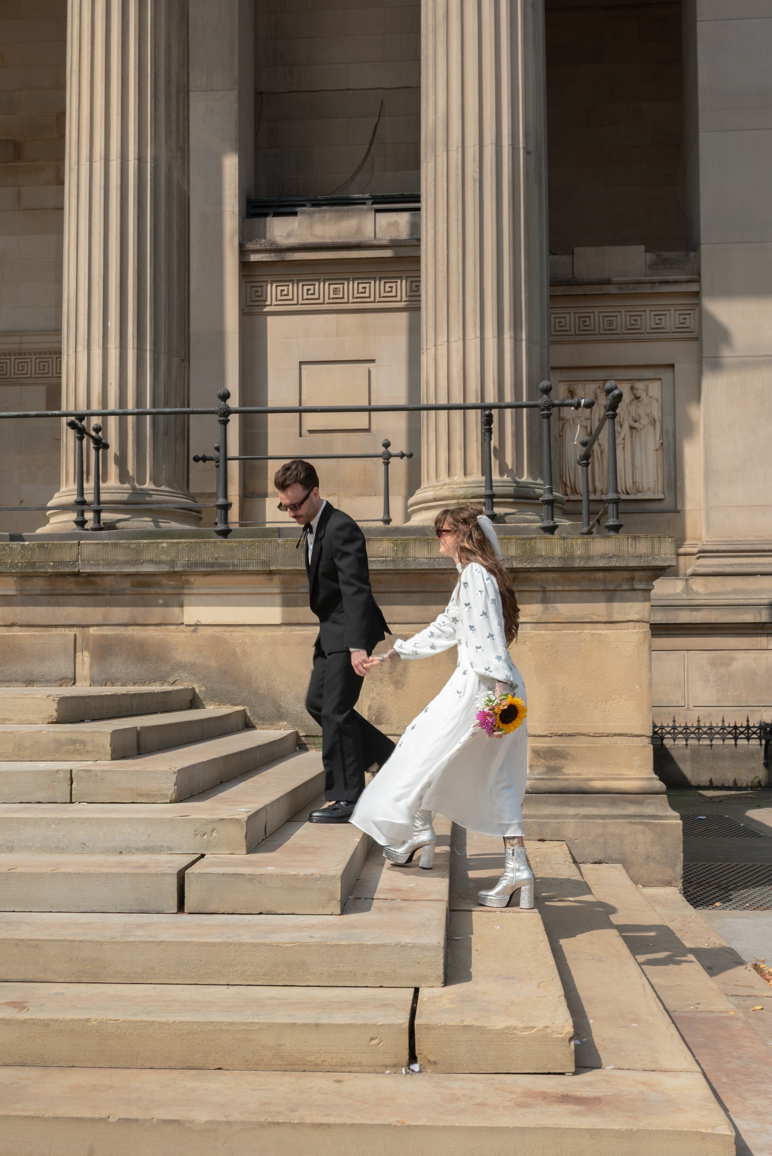 A young couple, dressed in wedding attire, holding hands and walking down stone steps outside a classical building with columns and ornate architectural details. The woman is wearing a white dress and silver platform boots, holding a bouquet of flowe