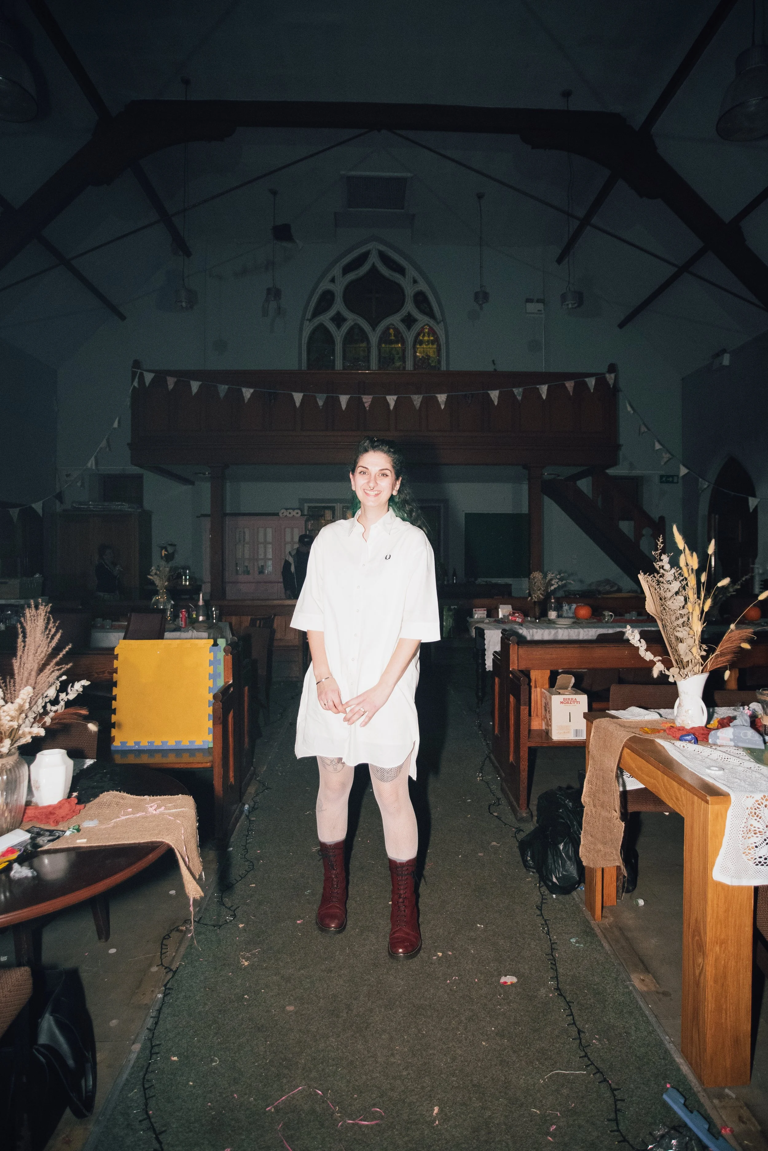 A woman with dark hair smiling, wearing a white oversized shirt, white fishnet stockings, and red lace-up boots, standing in the center of a decorated indoor space with tables, dried floral arrangements, and bunting in the background.