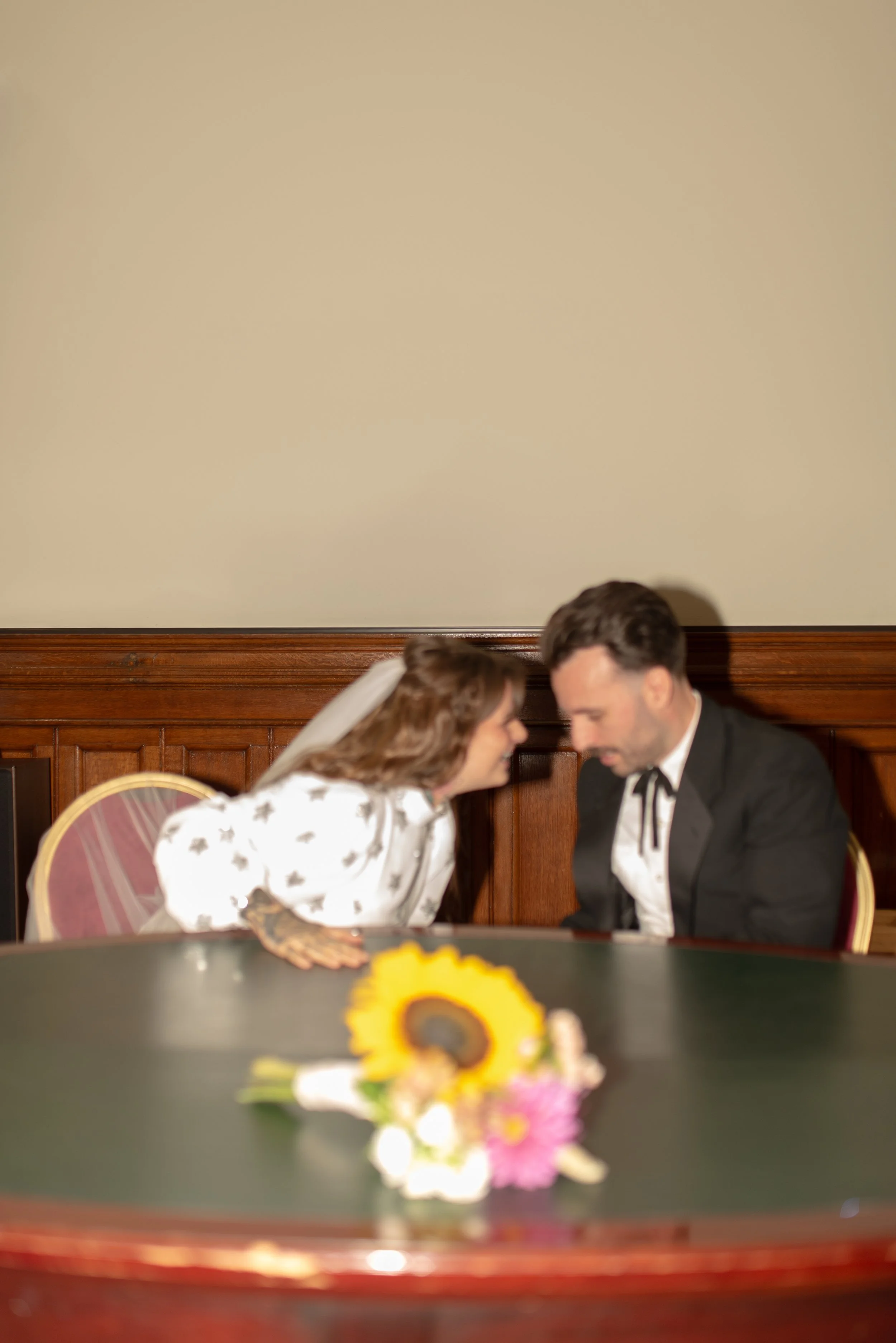 A bride and groom sitting close together at a wedding reception table with a sunflower centerpiece, leaning in towards each other with noses touching.