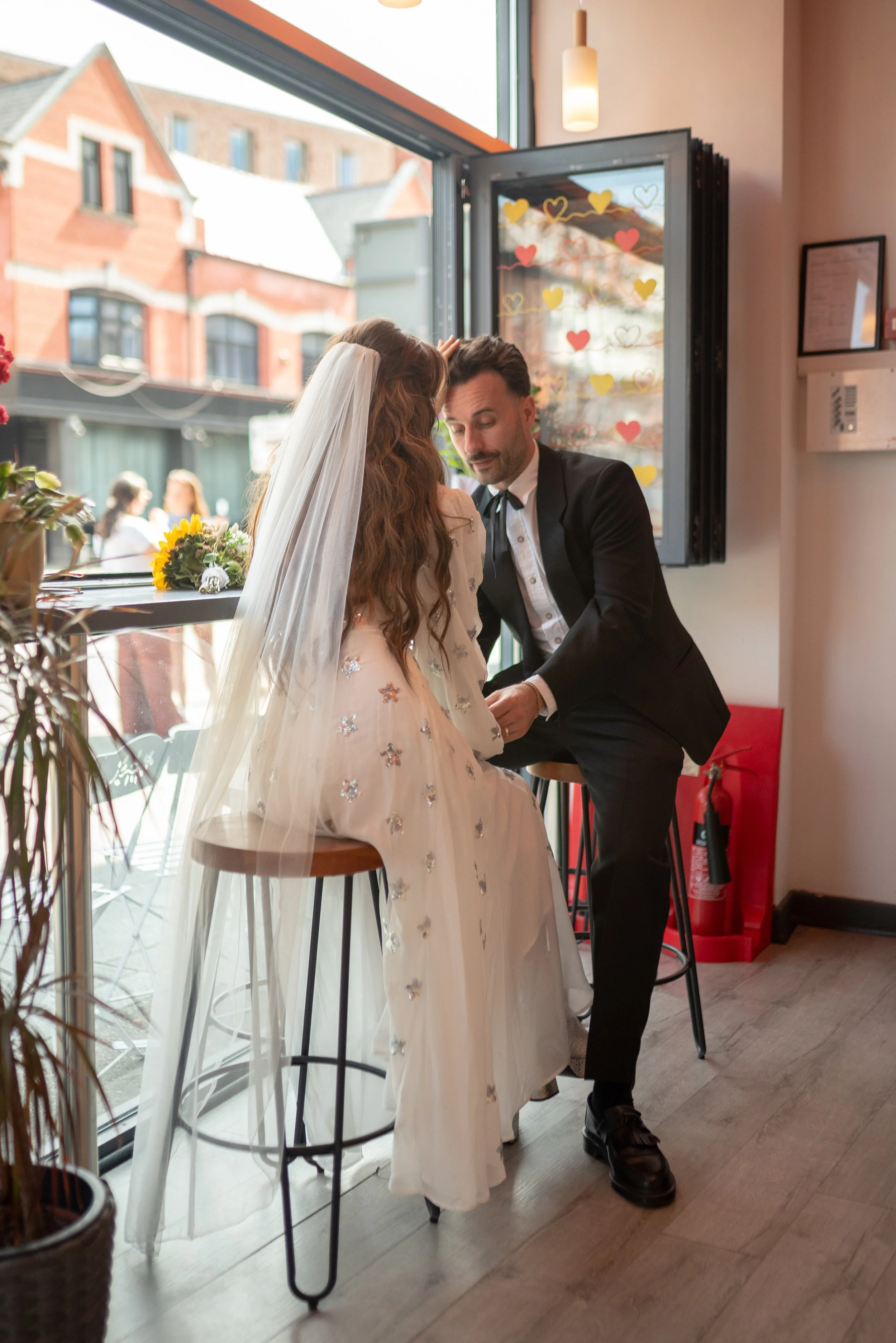 A bride with long, wavy hair wearing a white dress with floral embroidery and a veil sits on a stool inside a cafe, while a groom in a black tuxedo with a white shirt kneels in front of her, holding her hand. The scene is bright with large windows an