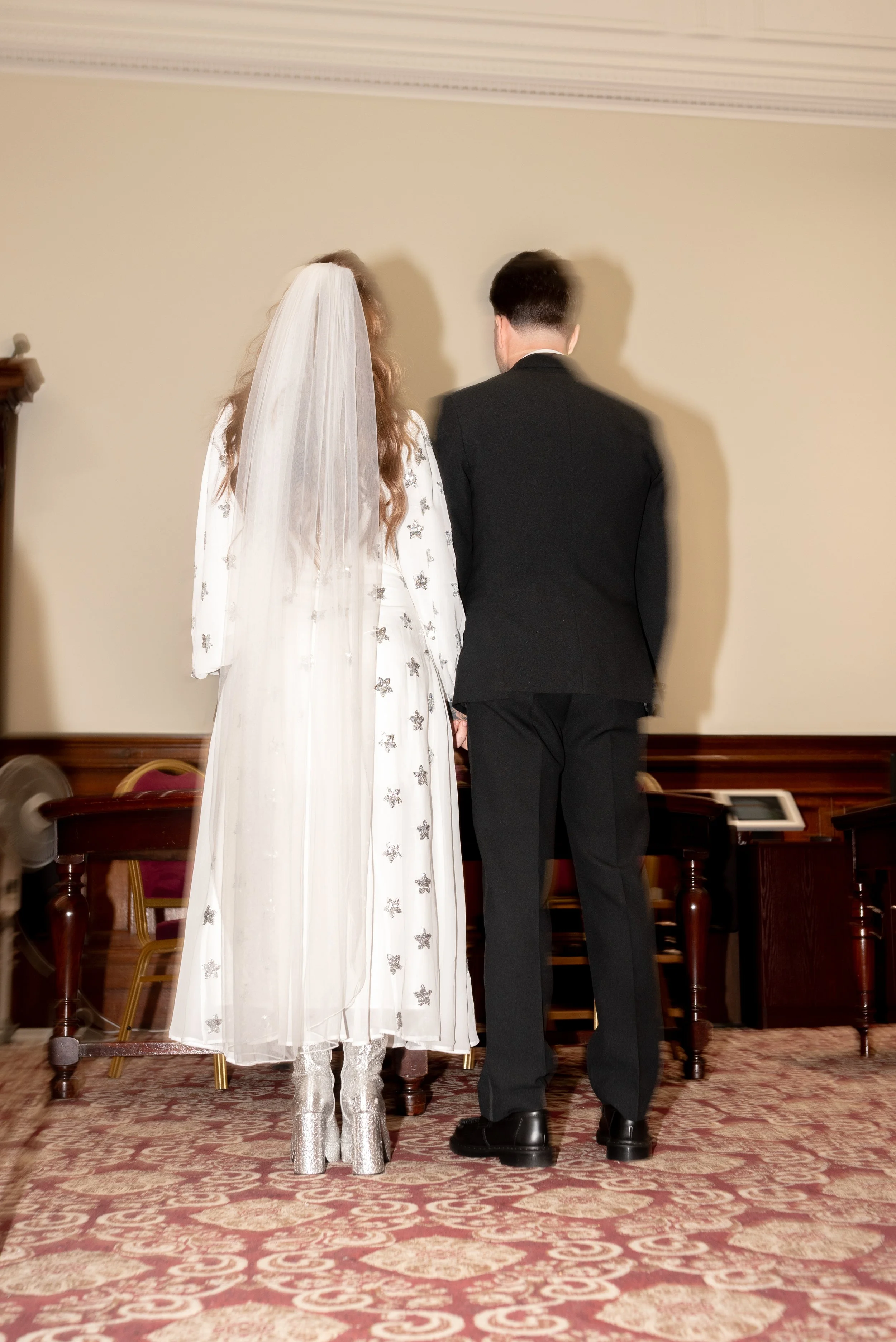 Bride and groom standing hand in hand with backs facing camera indoors during wedding ceremony.
