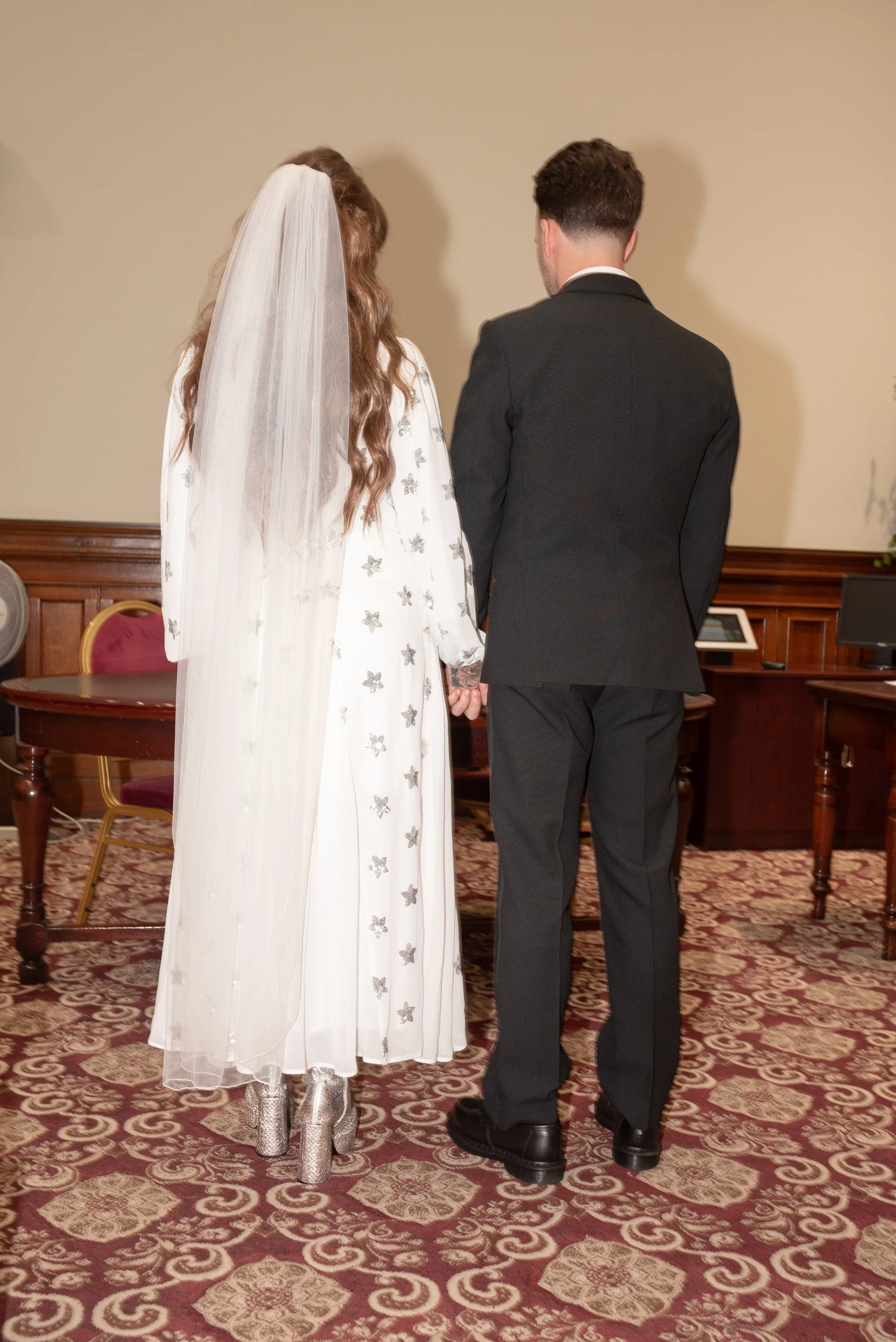 A bride and groom standing together holding hands with their backs facing the camera during their wedding ceremony.