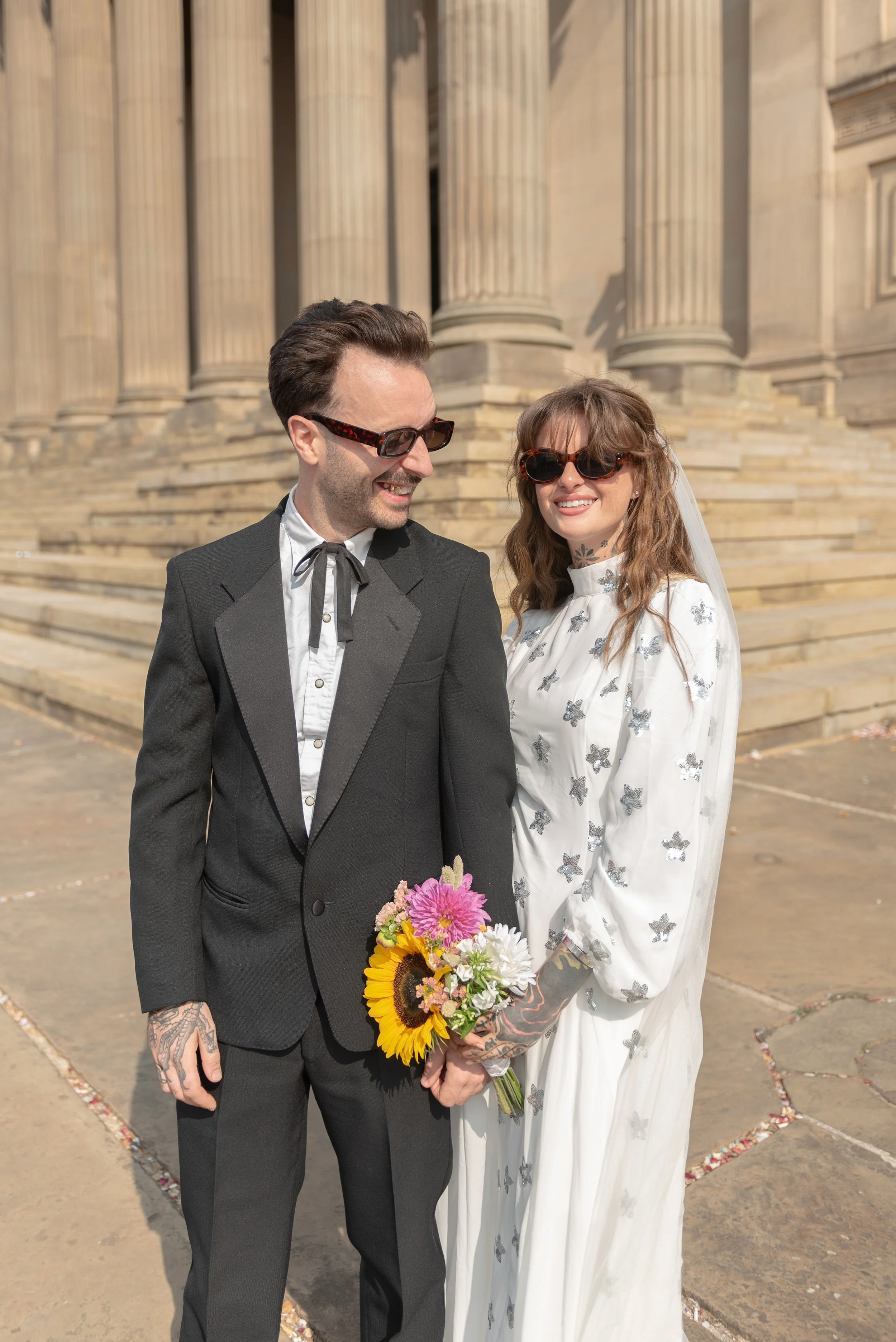 A couple dressed in formal wedding attire standing in front of a stone building, holding a bouquet of flowers, smiling and wearing sunglasses.