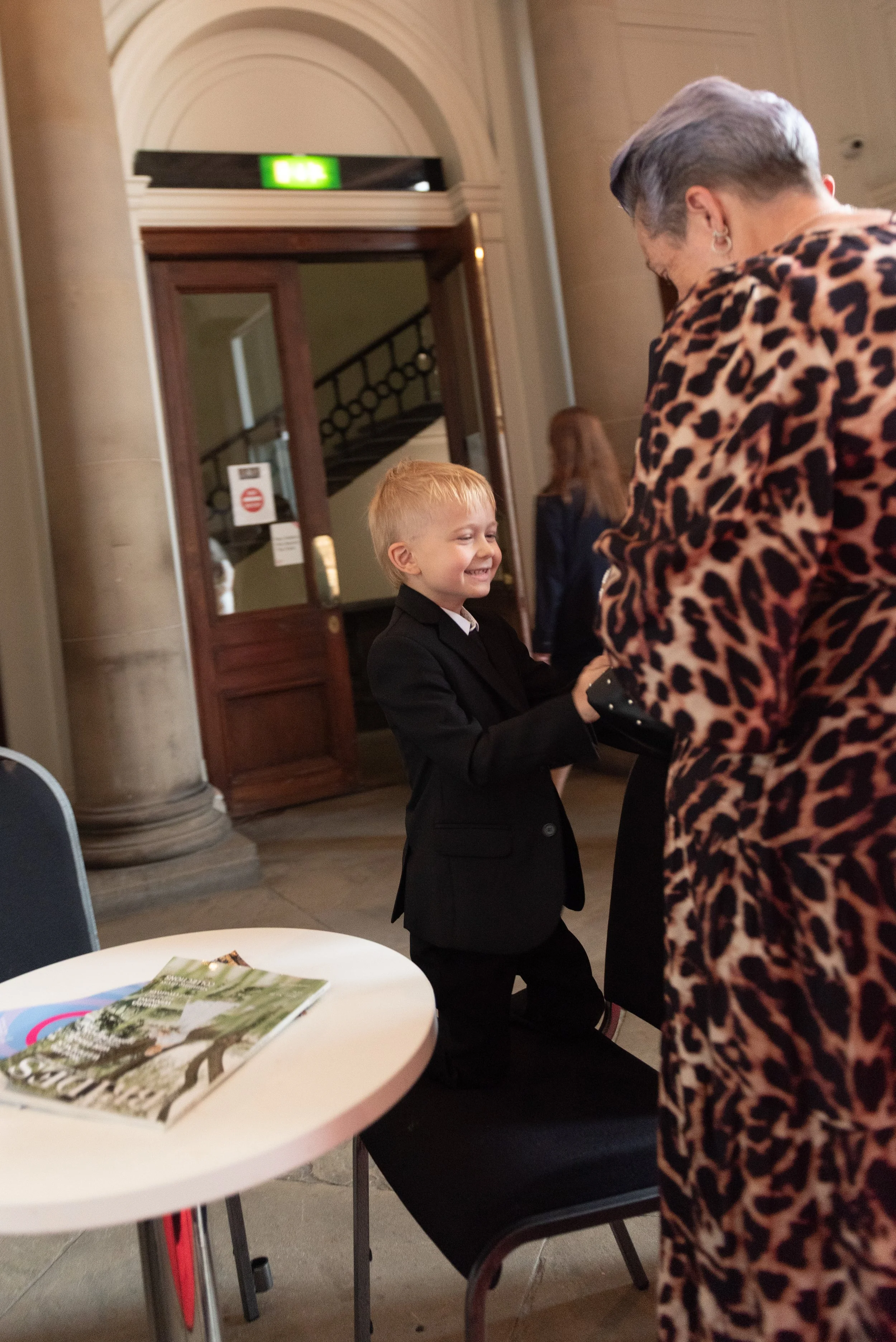 A young boy in a black suit kneeling on a chair, smiling as he exchanges a handshake with an older woman wearing a leopard print dress inside a building with tall columns and wooden doors.