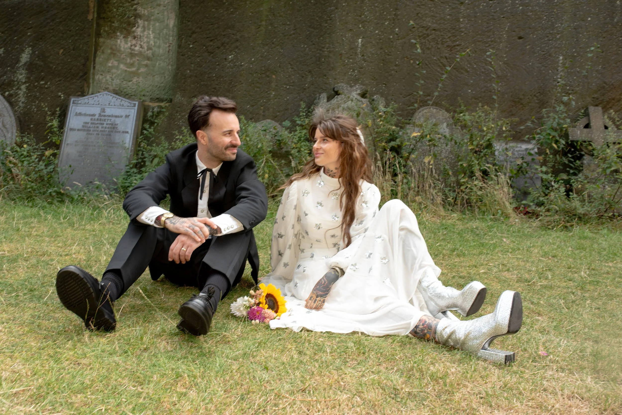 A man and woman sitting on the grass, looking at each other, with tombstones and greenery in the background.
