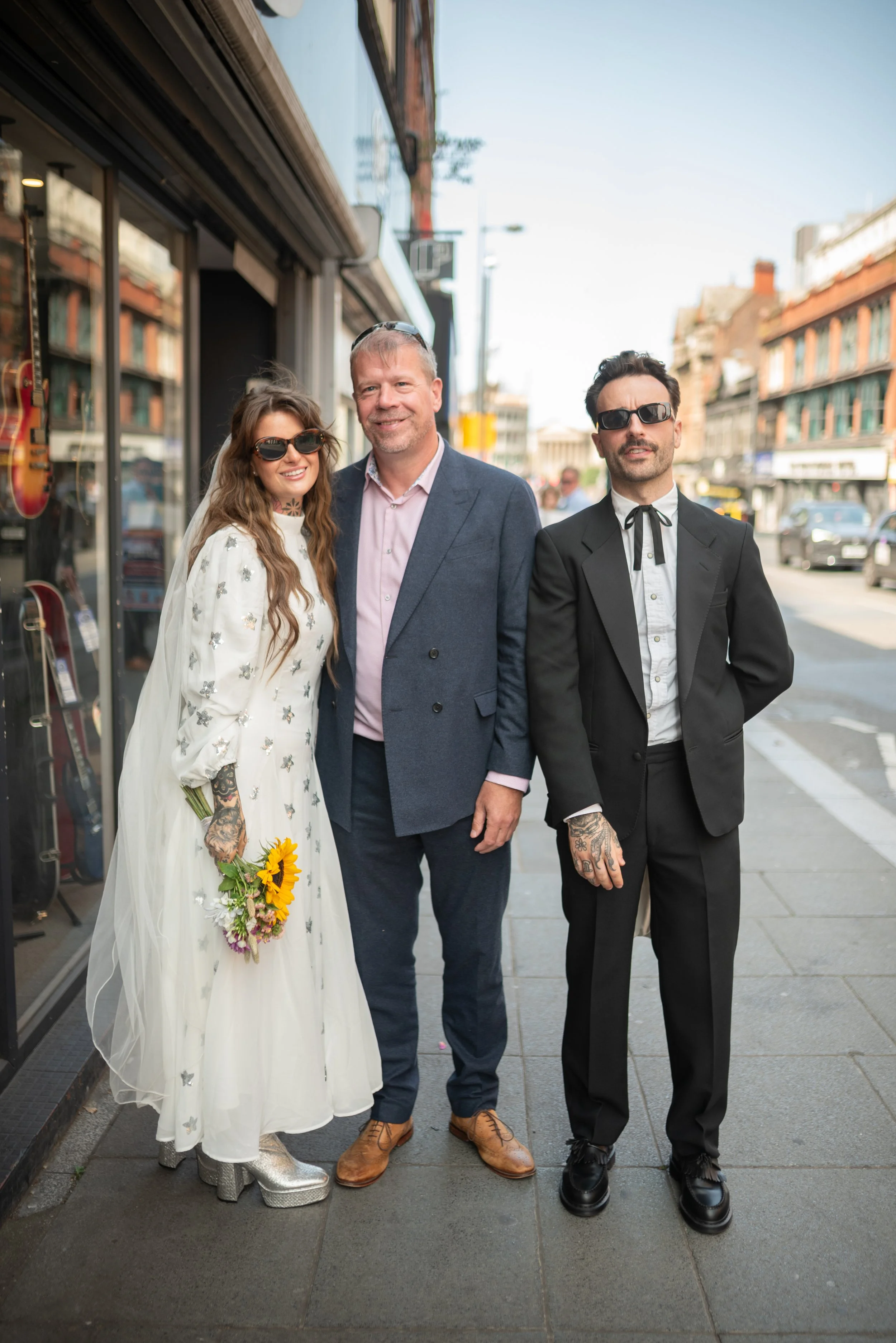 Three people pose on a city sidewalk, two men and a woman, dressed in stylish, semi-formal attire, with buildings and parked cars in the background.