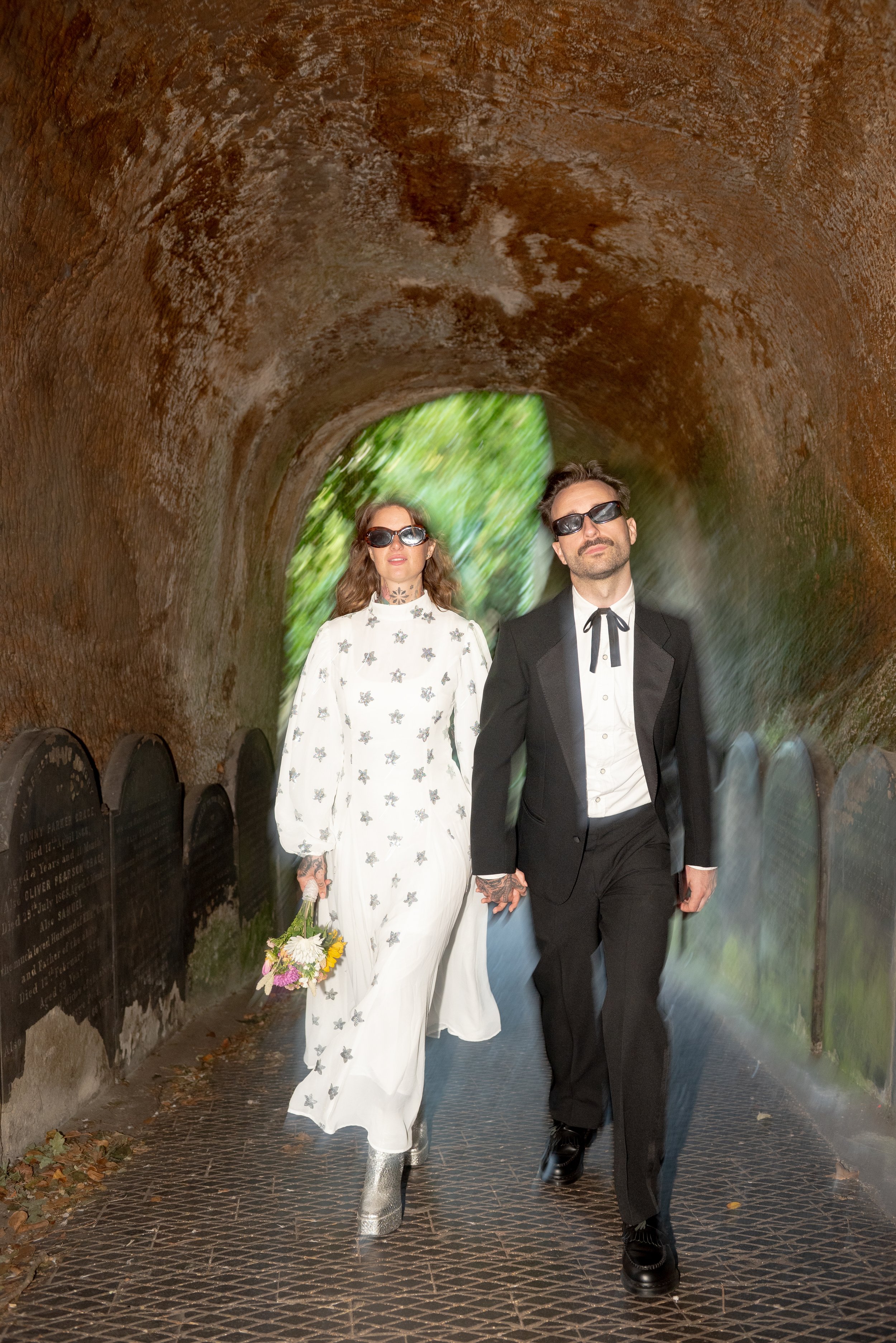 A man and woman, both wearing sunglasses and formal clothing, walk hand in hand through a tunnel with a textured, rusty brown ceiling and green foliage visible at the tunnel's exit. The woman is dressed in a white dress with floral patterns, holding 