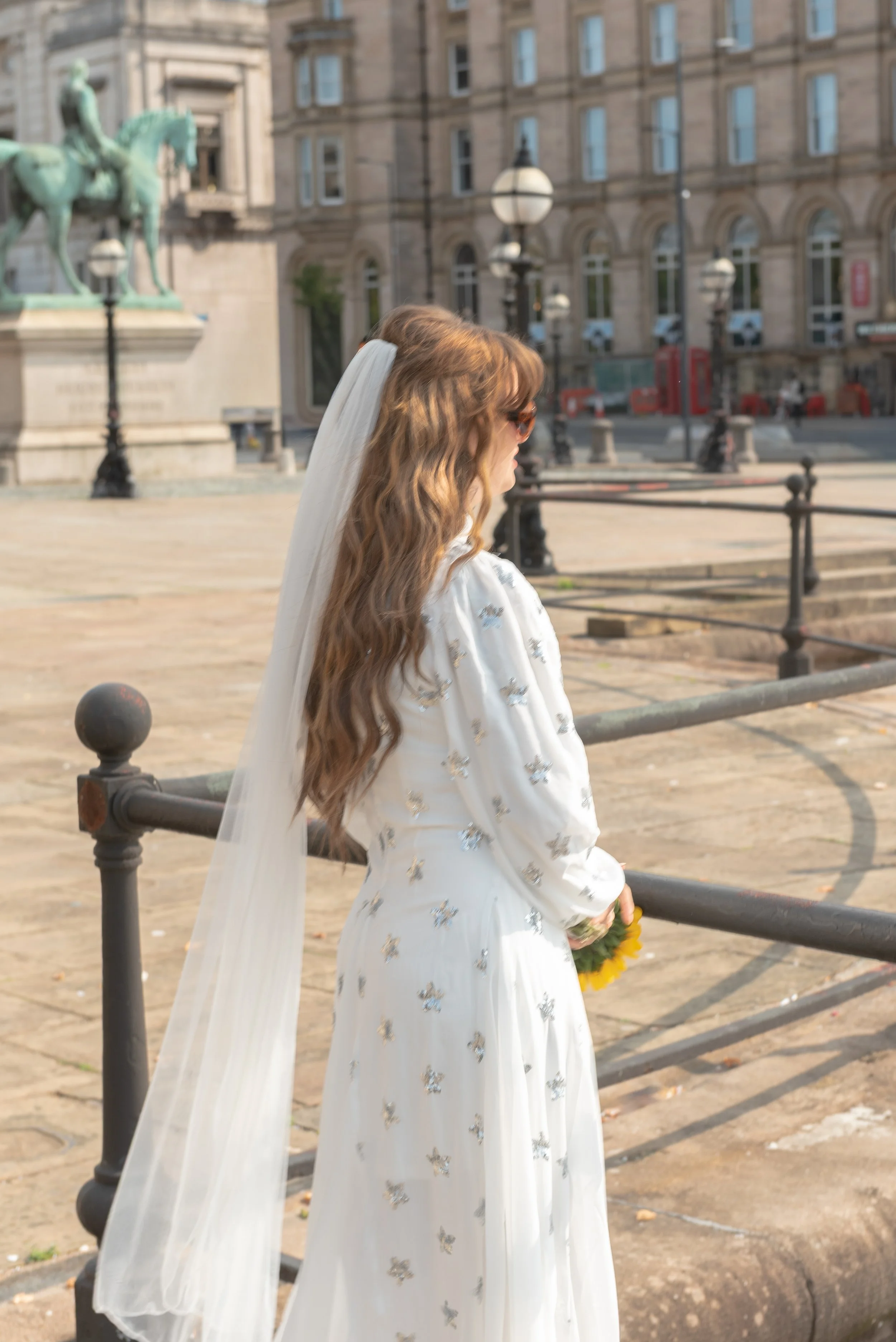 A woman with long wavy hair wearing a white dress with silver embellishments, a veil, and sunglasses standing outdoors near a metal fence, holding a sunflower, with a historic building and an equestrian statue in the background.