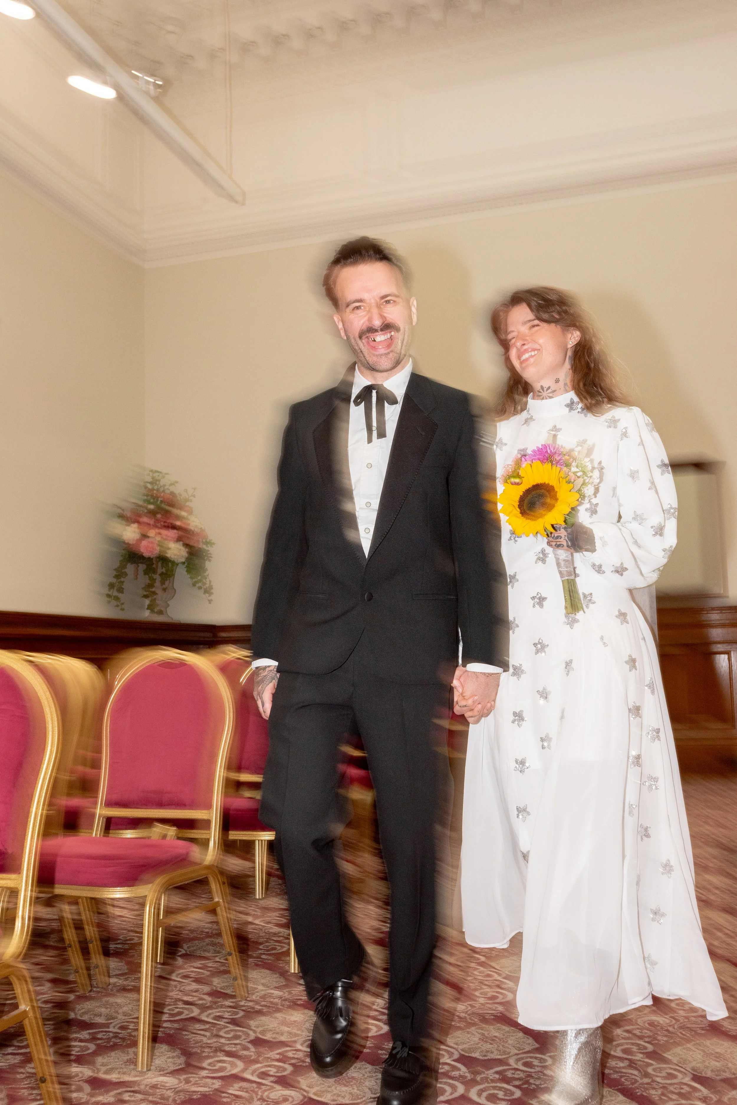 A couple holding hands, with the man wearing a black tuxedo and a woman in a white dress holding a sunflower bouquet, smiling at each other in a decorated room.