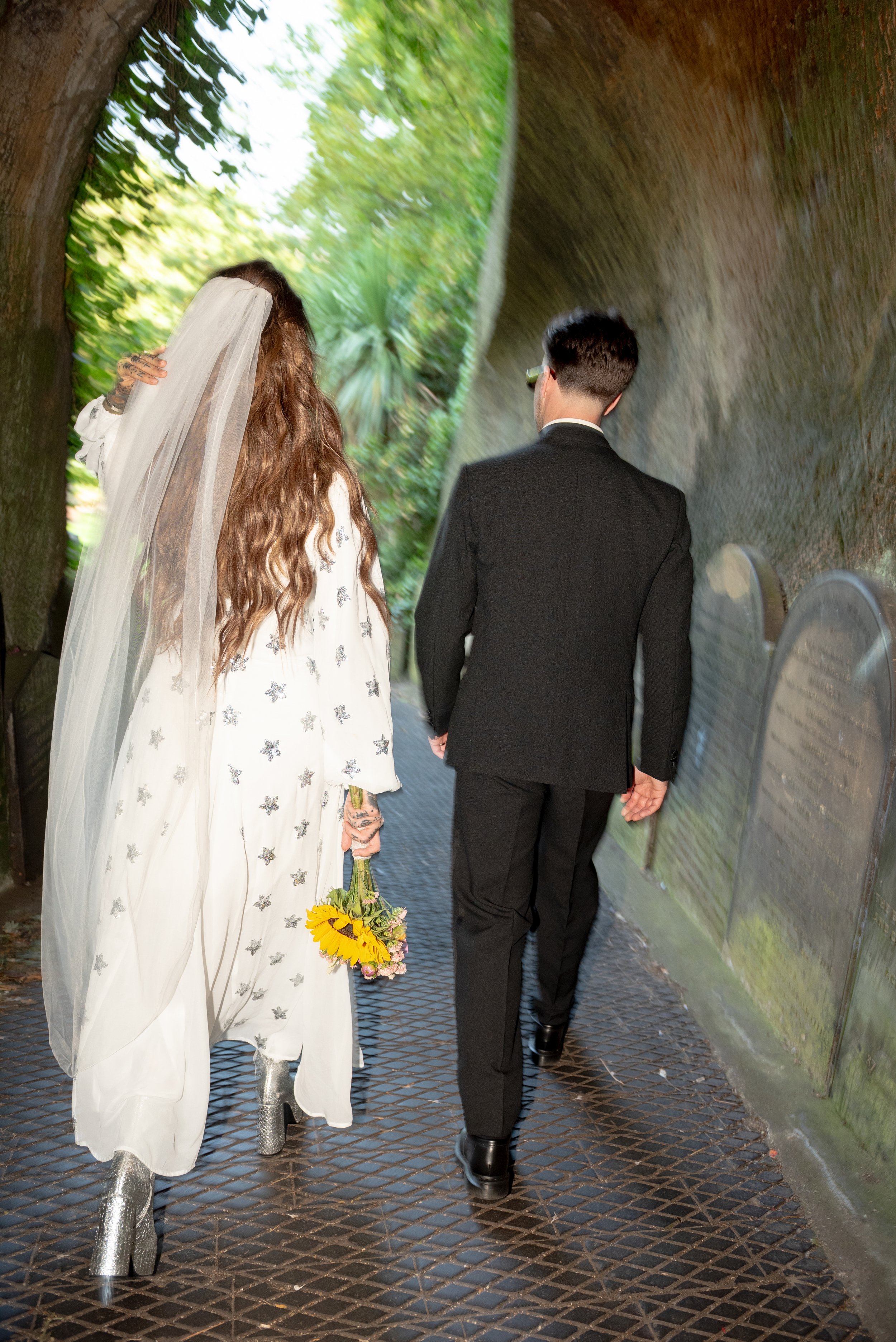 A couple walking outdoors in a tunnel-like area, the woman dressed in a white gown with star patterns and silver high-heeled boots, holding sunflowers and pink roses, with a veil over her wavy brown hair; the man dressed in a black suit with short ha