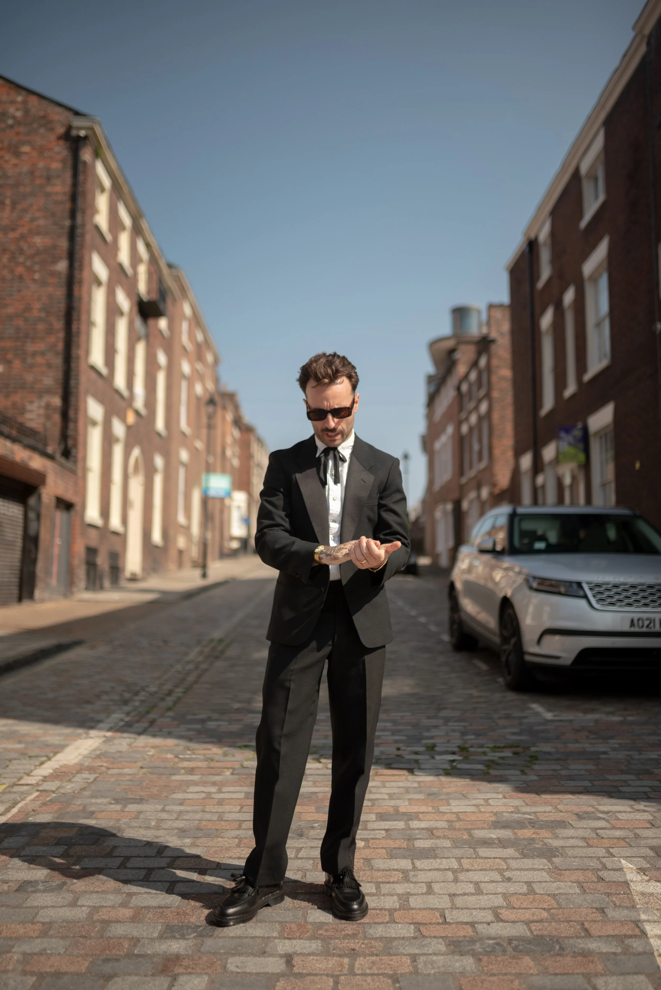 A man in a black tuxedo and sunglasses checking his watch on a cobblestone street with brick buildings and a parked white car.