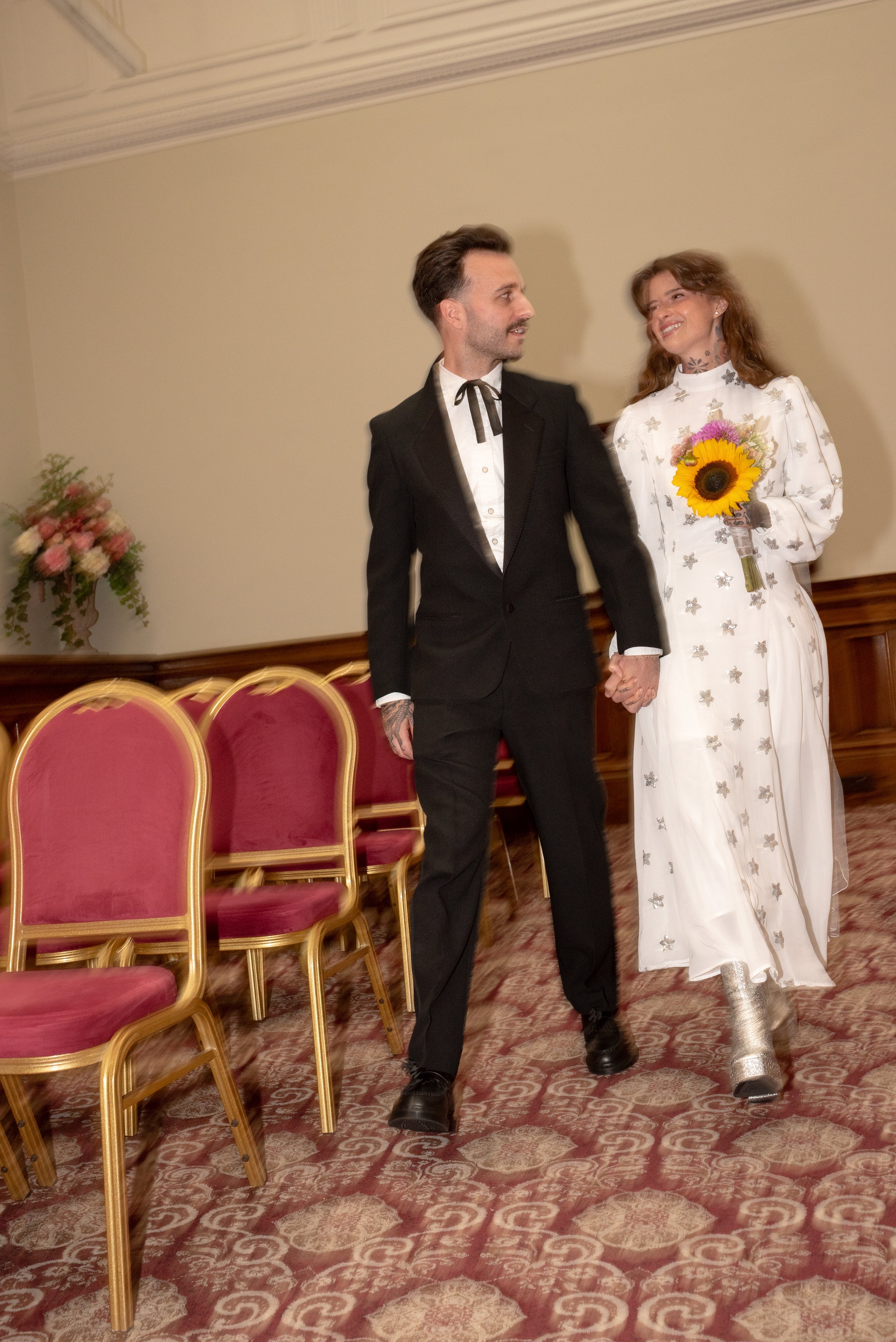 A couple, possibly at a wedding, holding hands and walking together in a room with red chairs and floral arrangements. The woman is smiling, holding a sunflower bouquet, and wearing a white dress with silver boots. The man is dressed in a black suit 