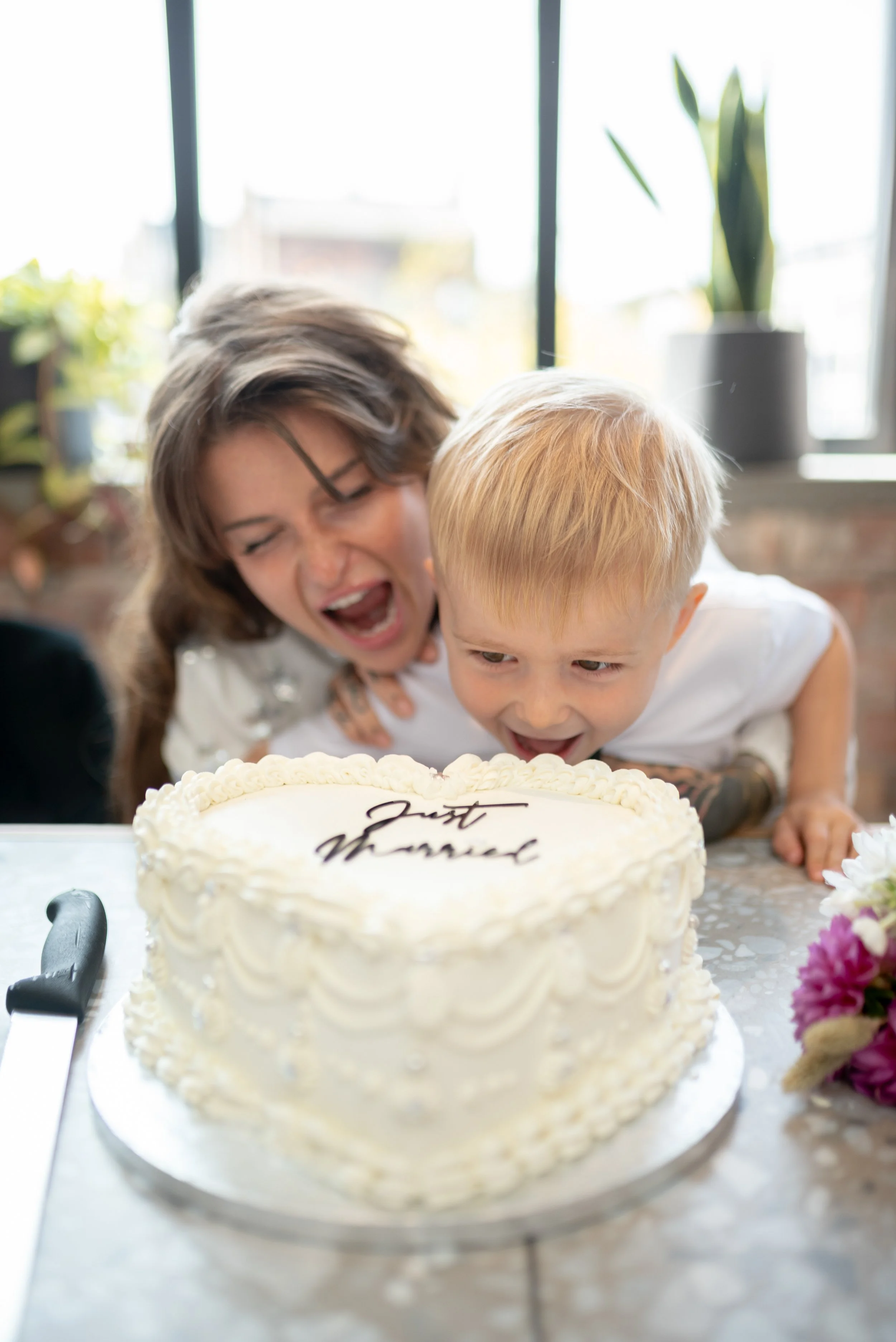 A woman and a young boy leaning over a birthday cake decorated with white frosting and a message that says "Just Married." The woman has short brown hair and is wearing glasses, while the boy has blonde hair and tattoos on his arms. They are celebrat