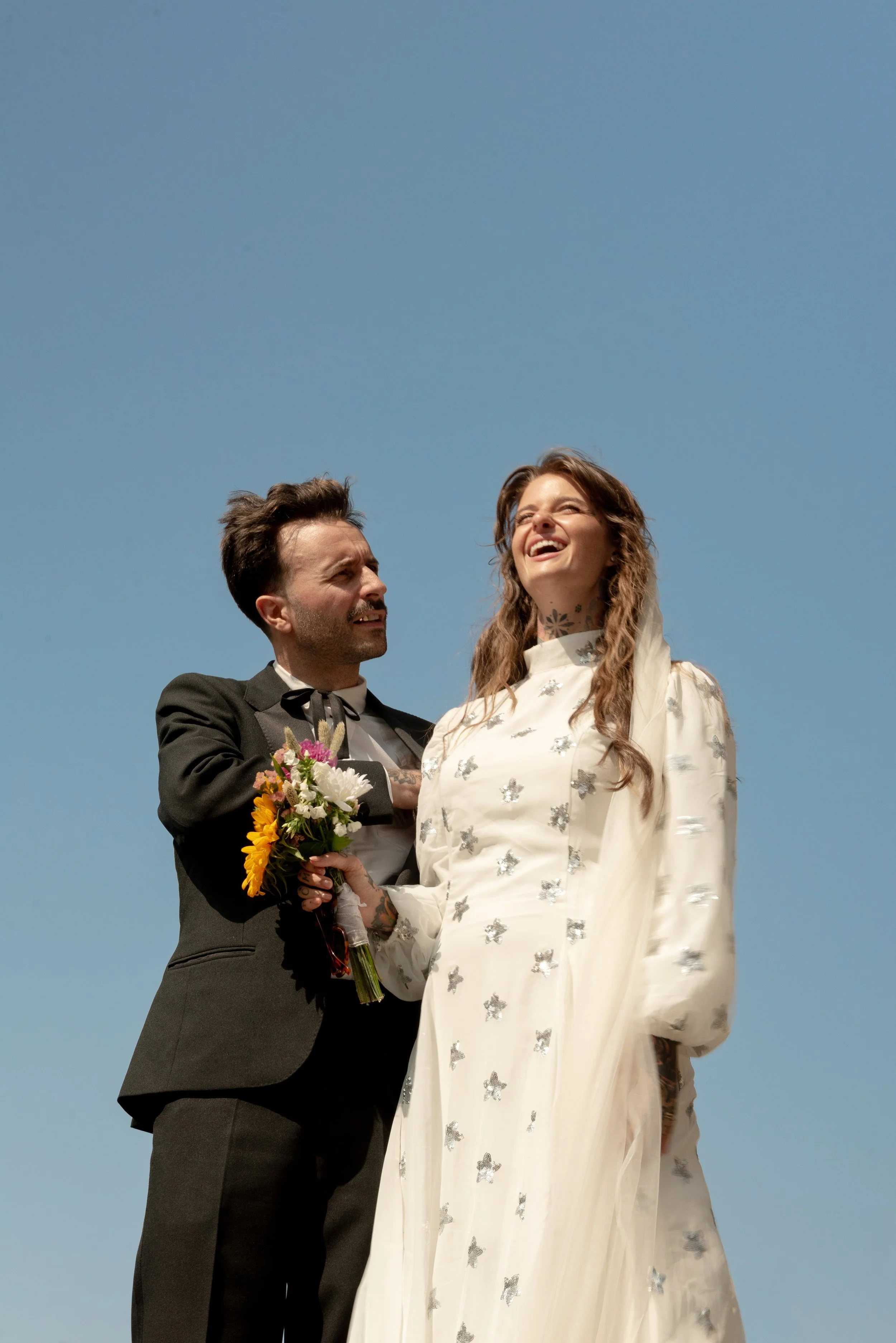 A groom in a black tuxedo holding a bouquet of flowers stands next to a smiling bride in a white wedding dress with silver embellishments, under a clear blue sky.
