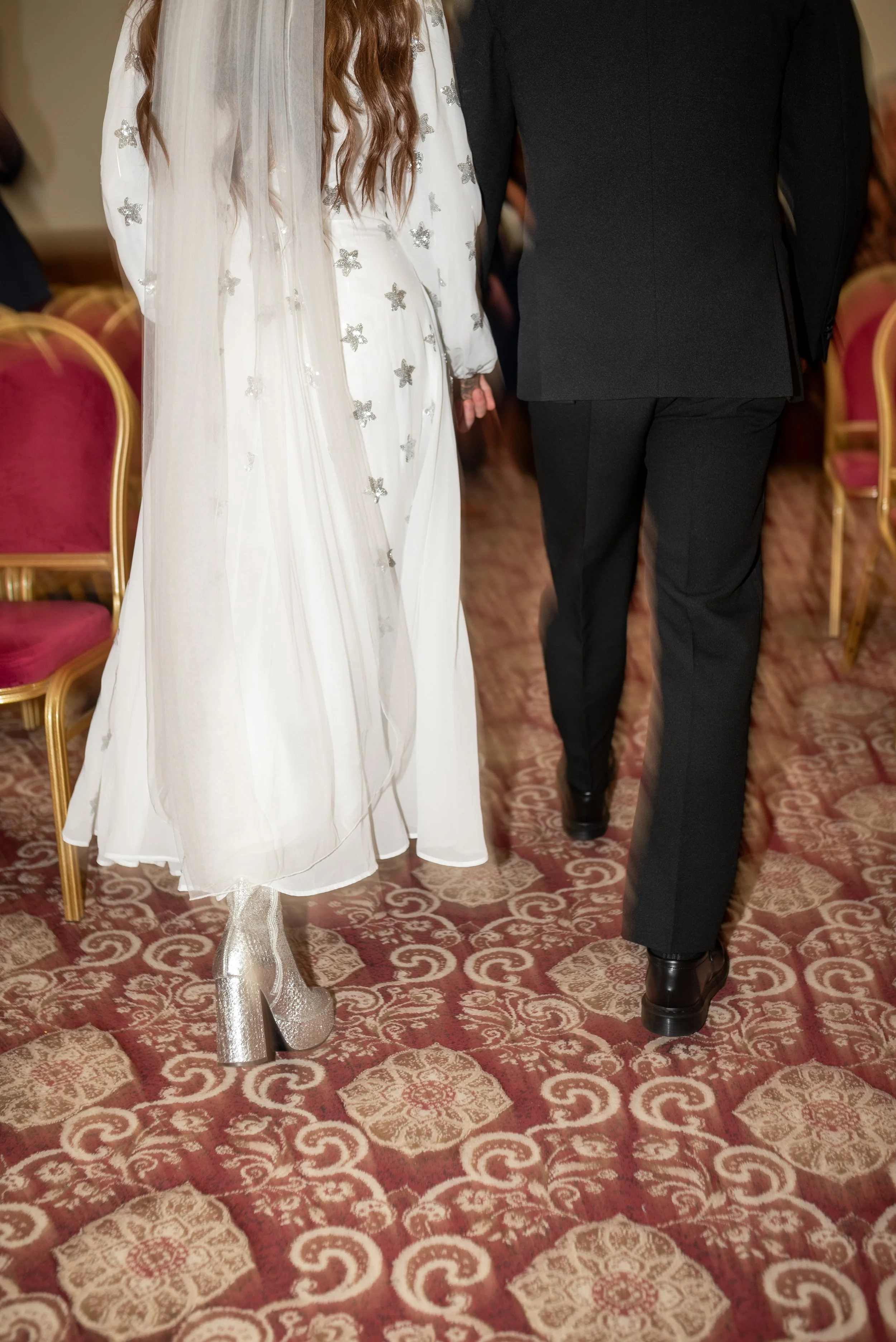 Couple dressed in formal attire, holding hands, walking on patterned carpet, with red velvet chairs in the background.