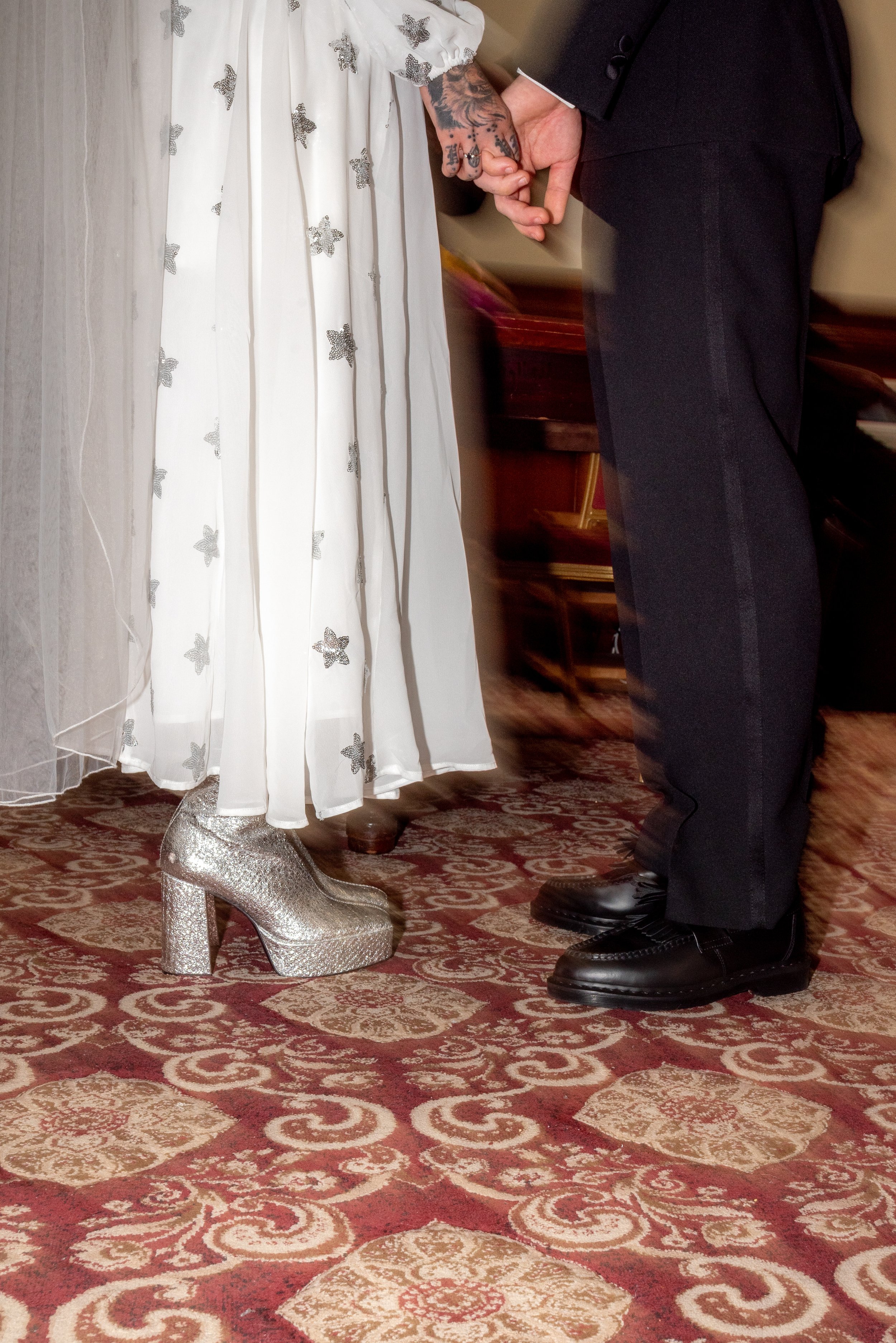 Close-up of a bride and groom holding hands, with the bride wearing a white dress with butterfly patterns and sparkly high heels, and the groom in black pants and shoes, standing on a patterned carpet.