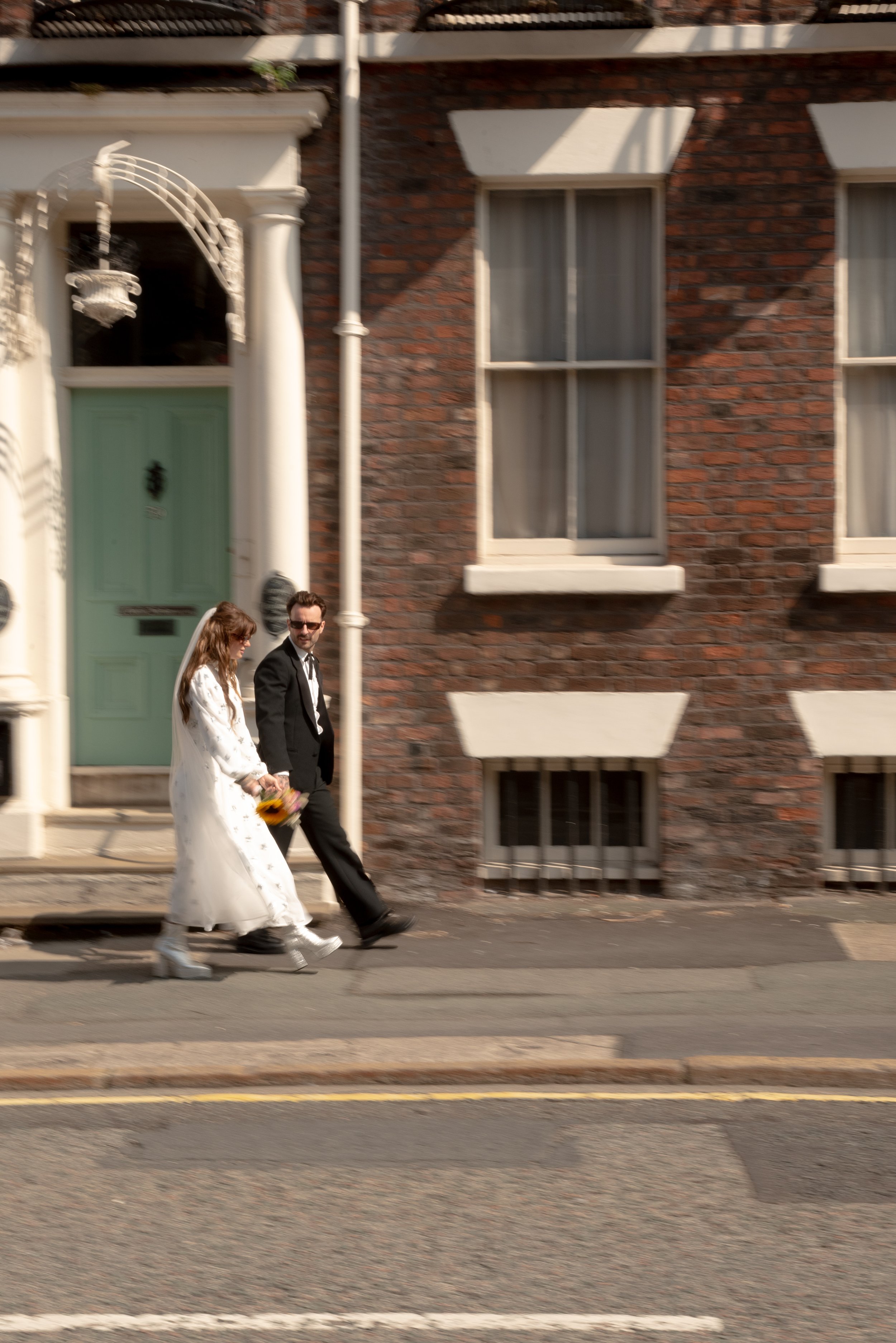 A bride and groom walking hand-in-hand on the sidewalk in front of a brick house, with the bride holding a bouquet of flowers and wearing a white dress, and the groom dressed in a black tuxedo.