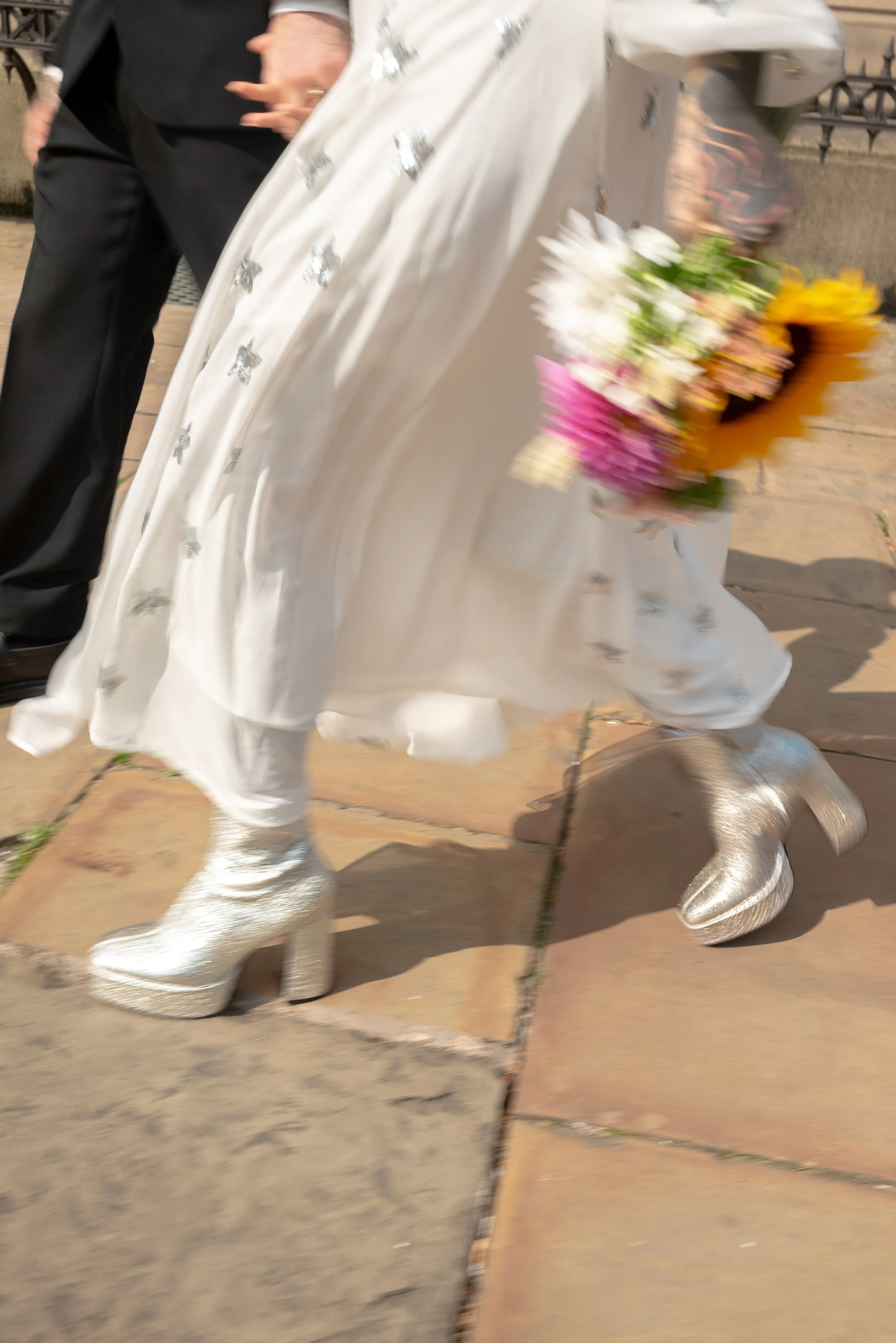 Person wearing shiny silver platform boots and a flowing white dress with silver embroidery, holding a colorful bouquet of flowers, walking on a tiled outdoor surface.