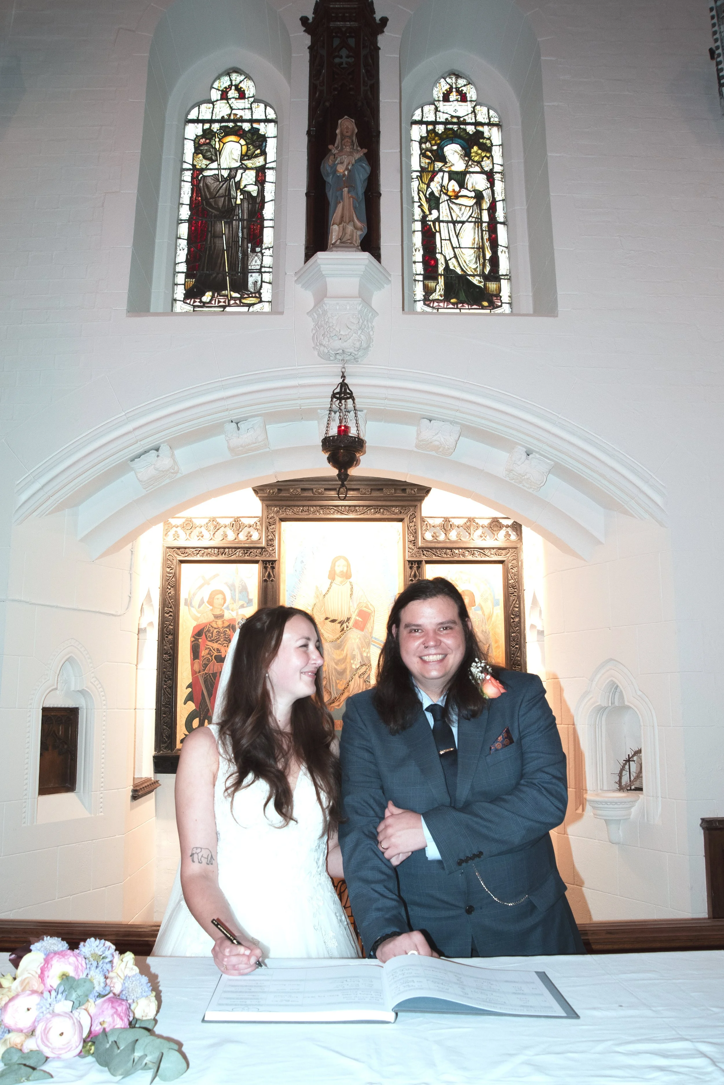 A couple stands at an altar inside a church, with the woman in a white wedding dress holding a pen and the man in a blue suit with a boutonniere, both smiling. The church features stained glass windows and religious artwork behind them.